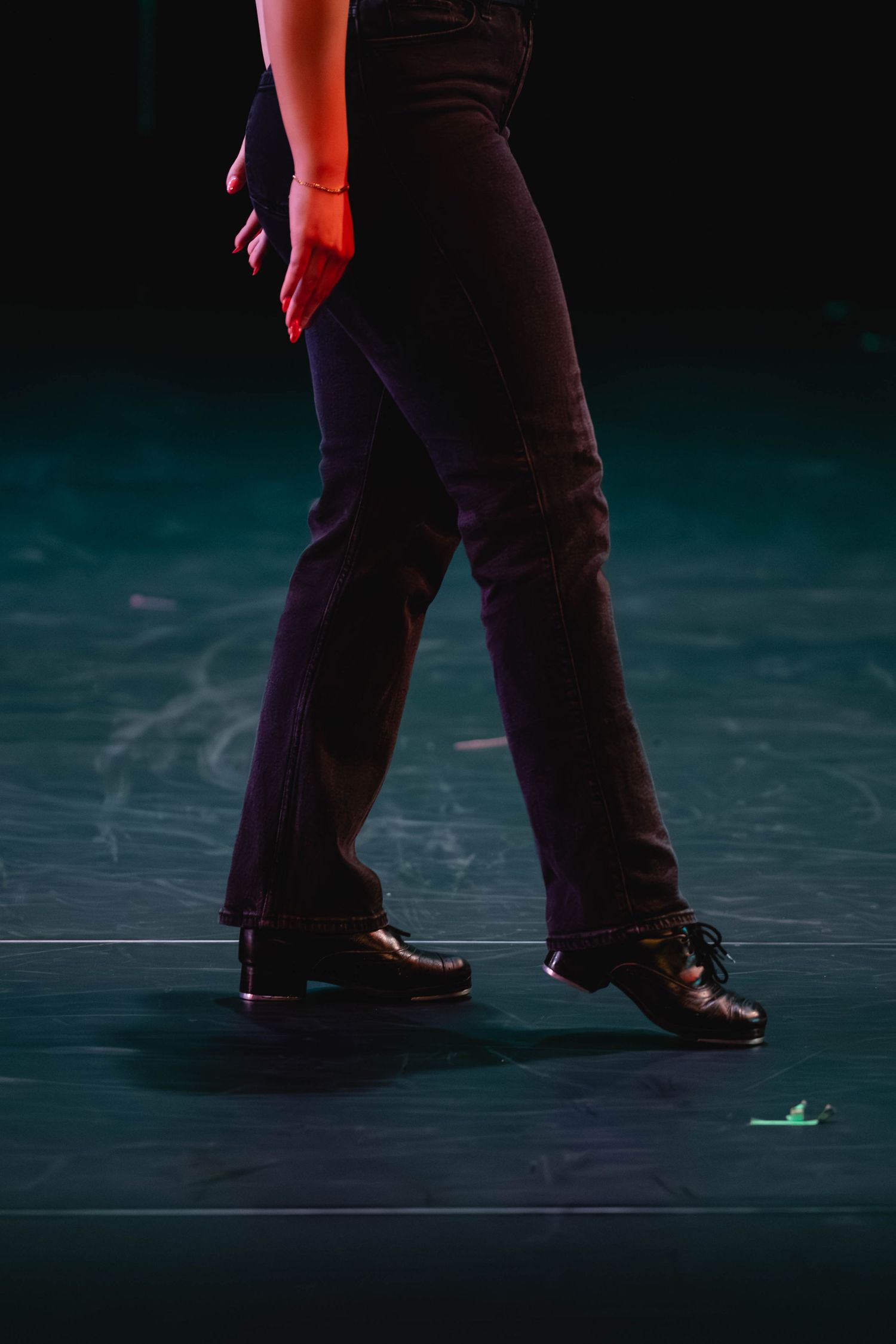 Close-up of a dancer's legs in black pants and tap shoes performing on a dark stage with dramatic lighting.