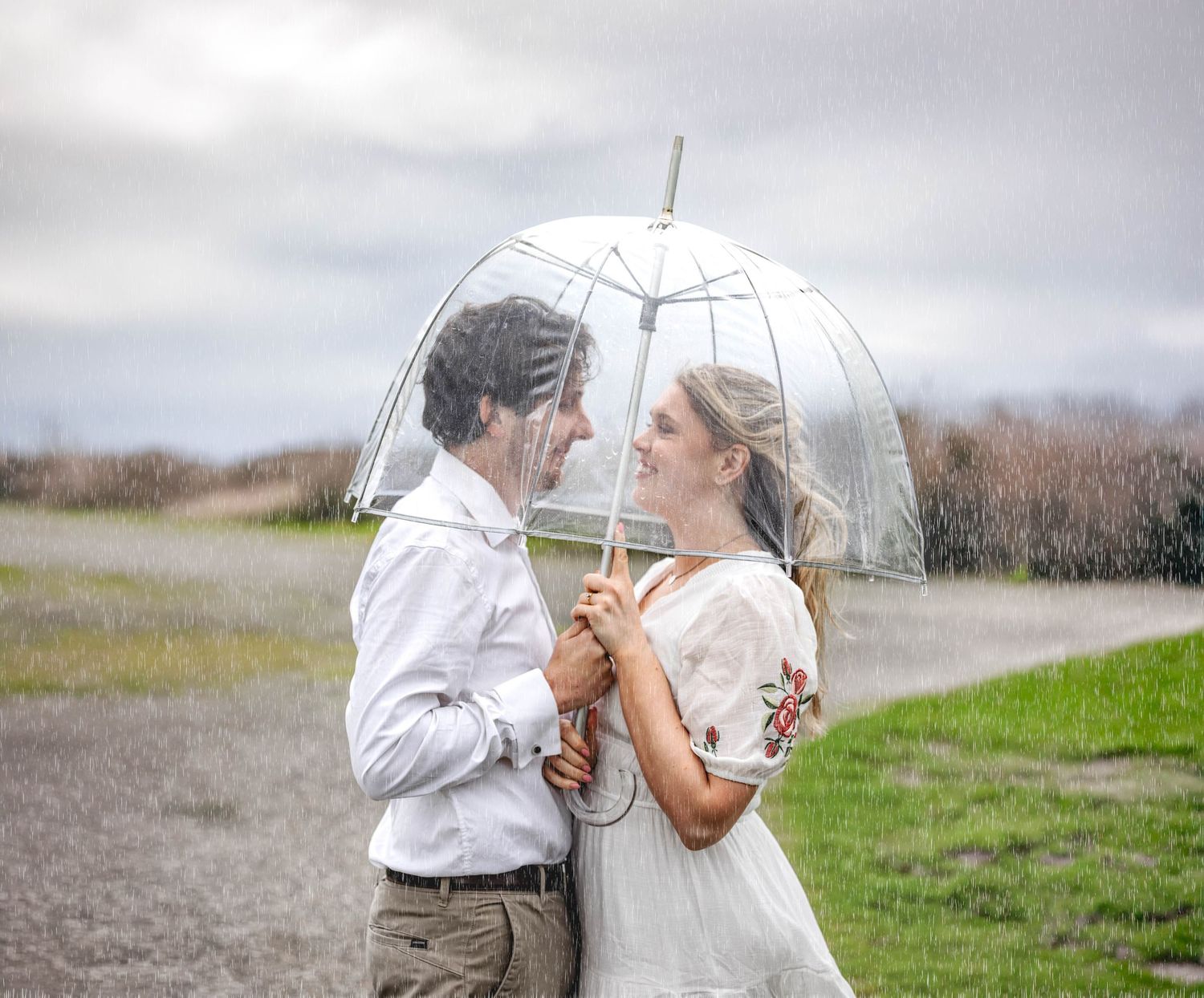 Couple sharing a clear bubble umbrella in the rain, smiling at each other in a romantic outdoor wedding photo.