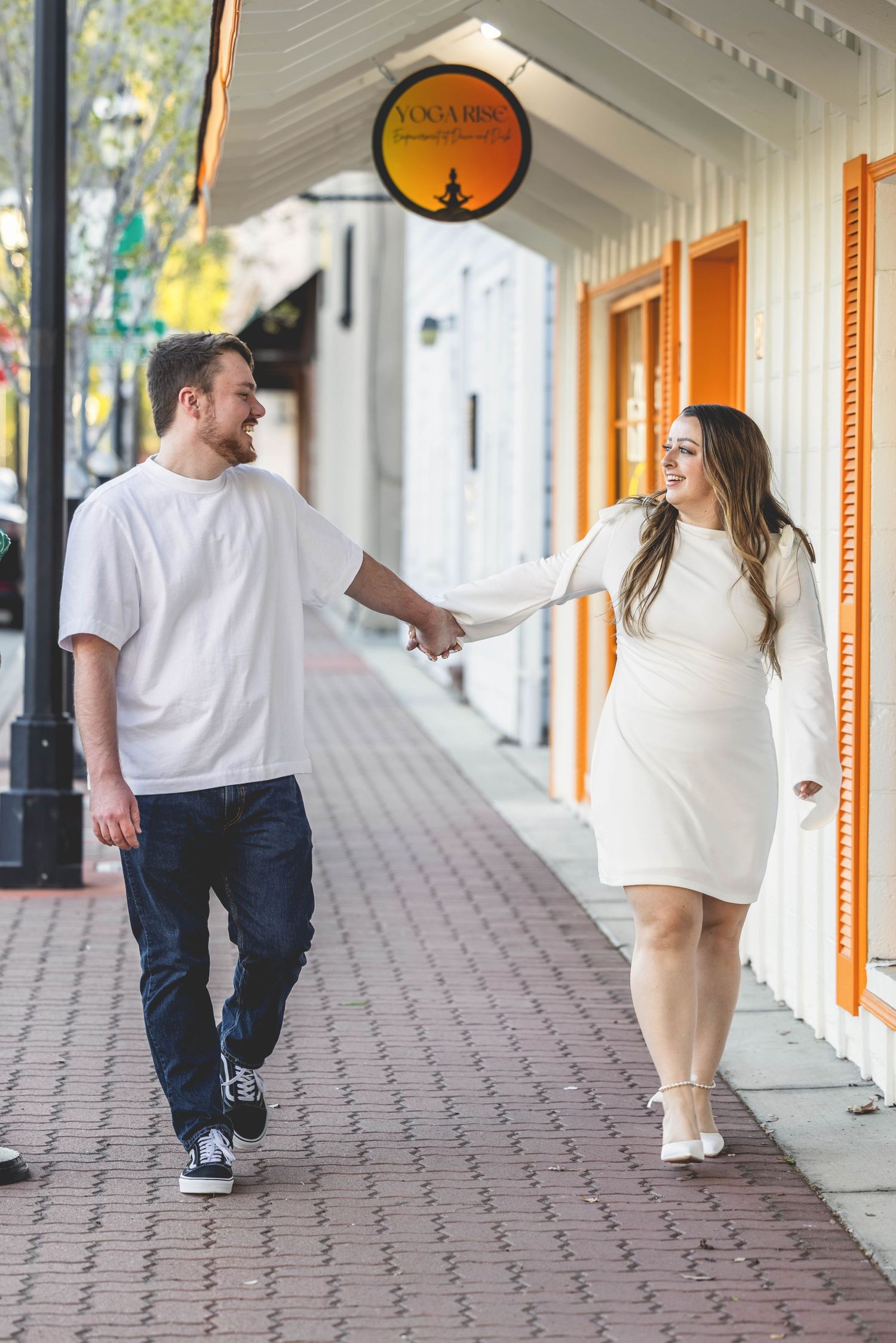 Couple holding hands walking on a brick sidewalk, both wearing white outfits, smiling at each other outside a yoga studio.