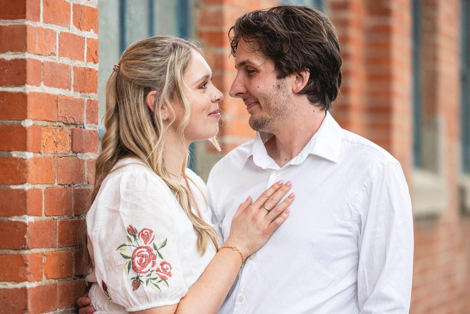 Couple sharing a romantic moment by a brick wall, woman in floral embroidered dress gazing at man in white shirt.