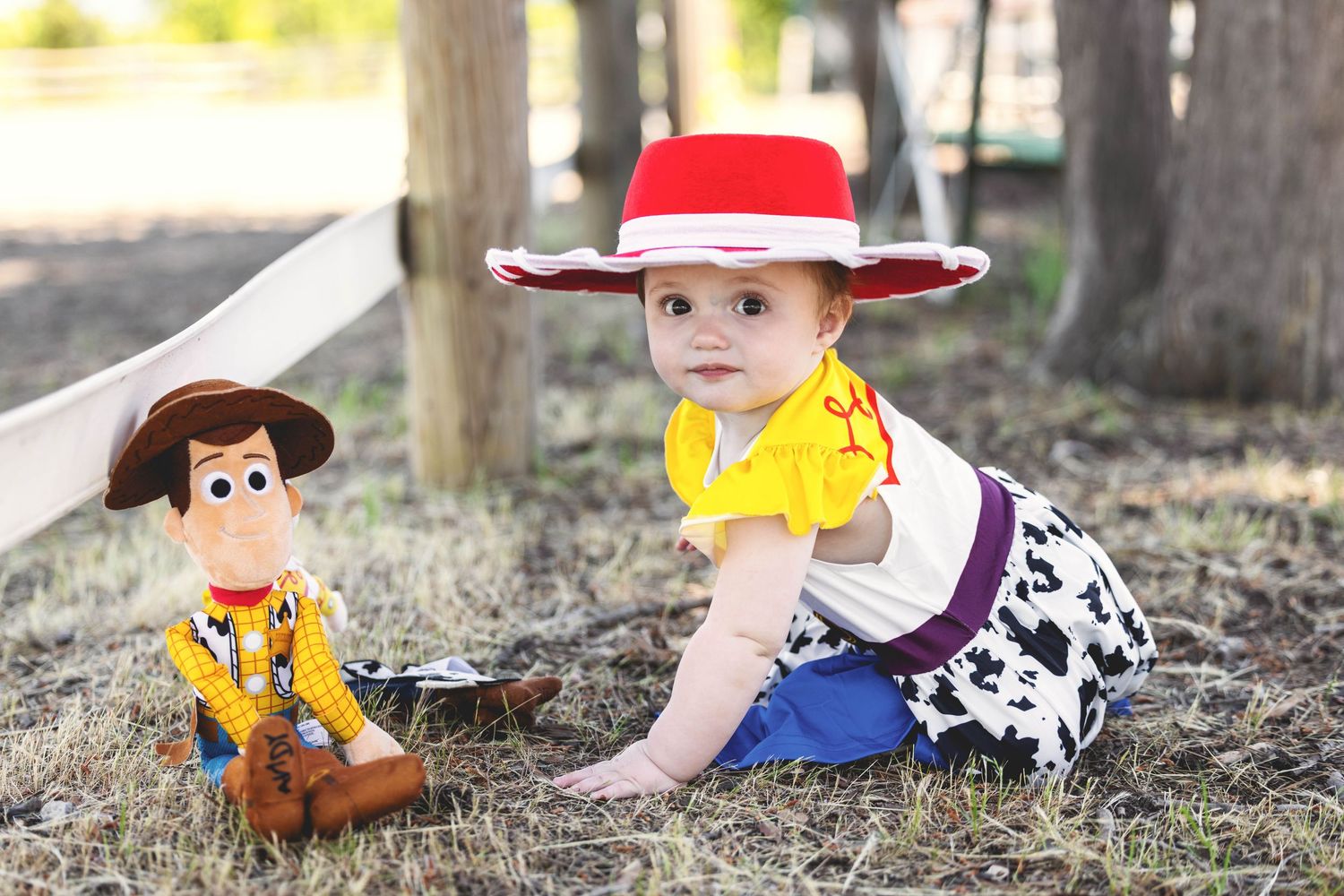 Baby dressed as Jessie from Toy Story sitting on grass next to a Woody doll outdoors.