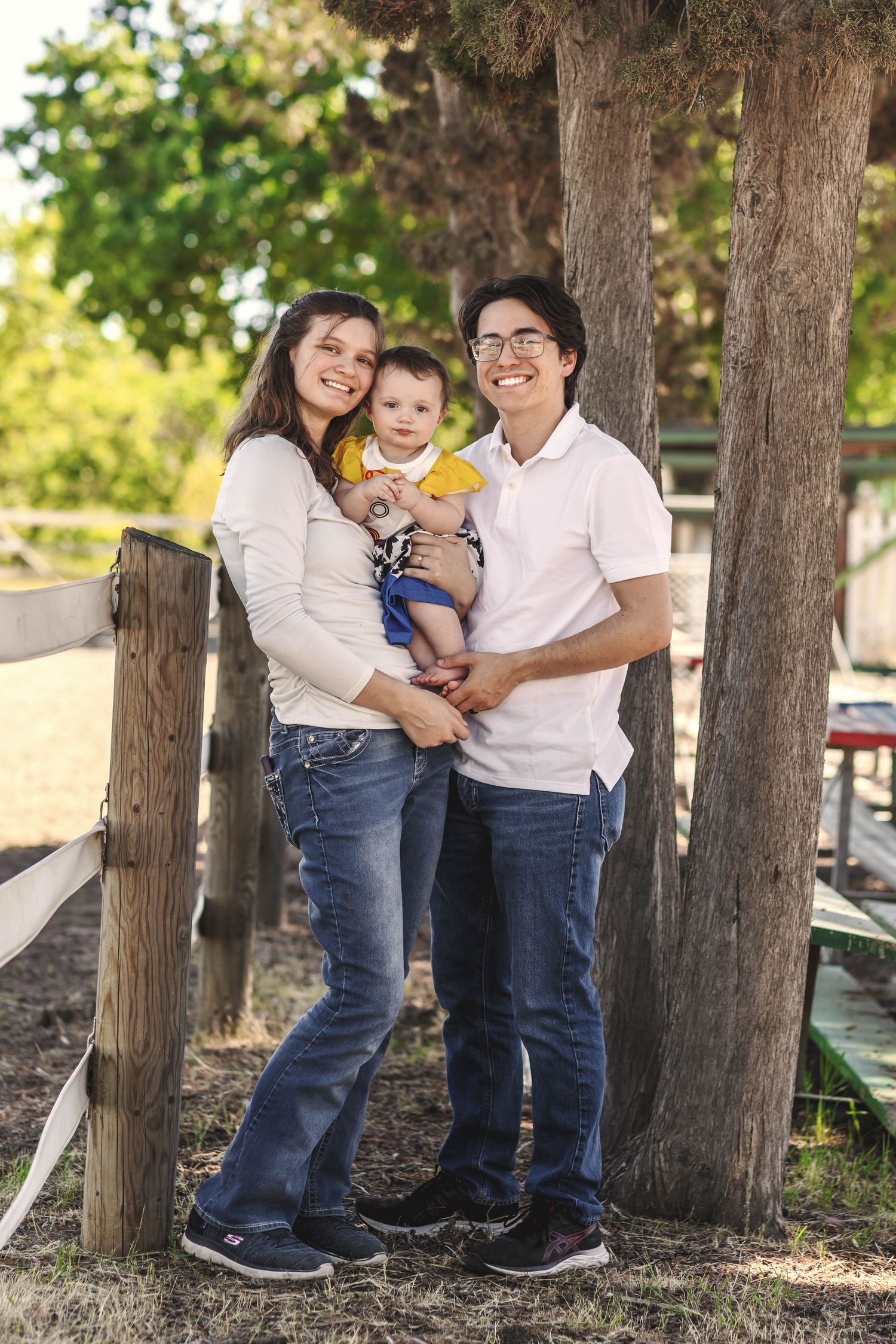 Happy young family with baby boy posing outdoors near wooden fence and trees on a sunny day.