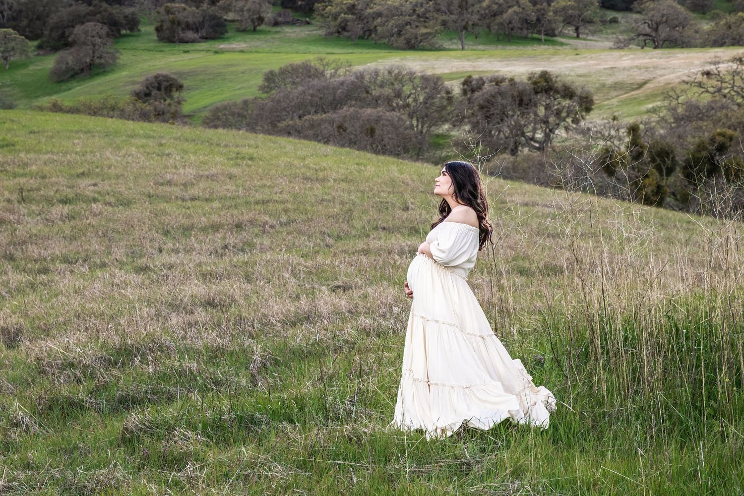 Pregnant woman in white dress standing in green rolling hills field during outdoor maternity photo session.