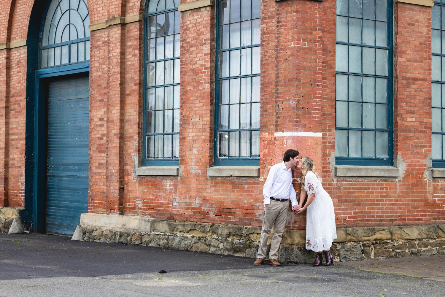 Couple sharing a romantic kiss against a rustic red brick building with teal windows during an outdoor engagement session.