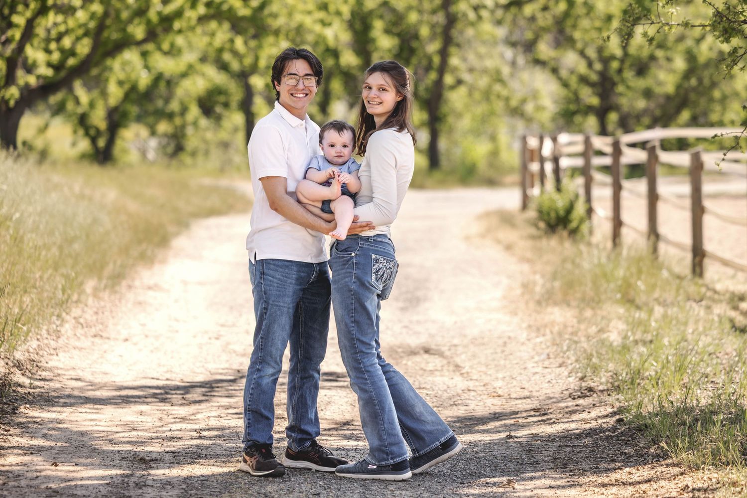 A young family of three poses on a sunny dirt path, parents smiling as mother holds baby, surrounded by lush green trees.