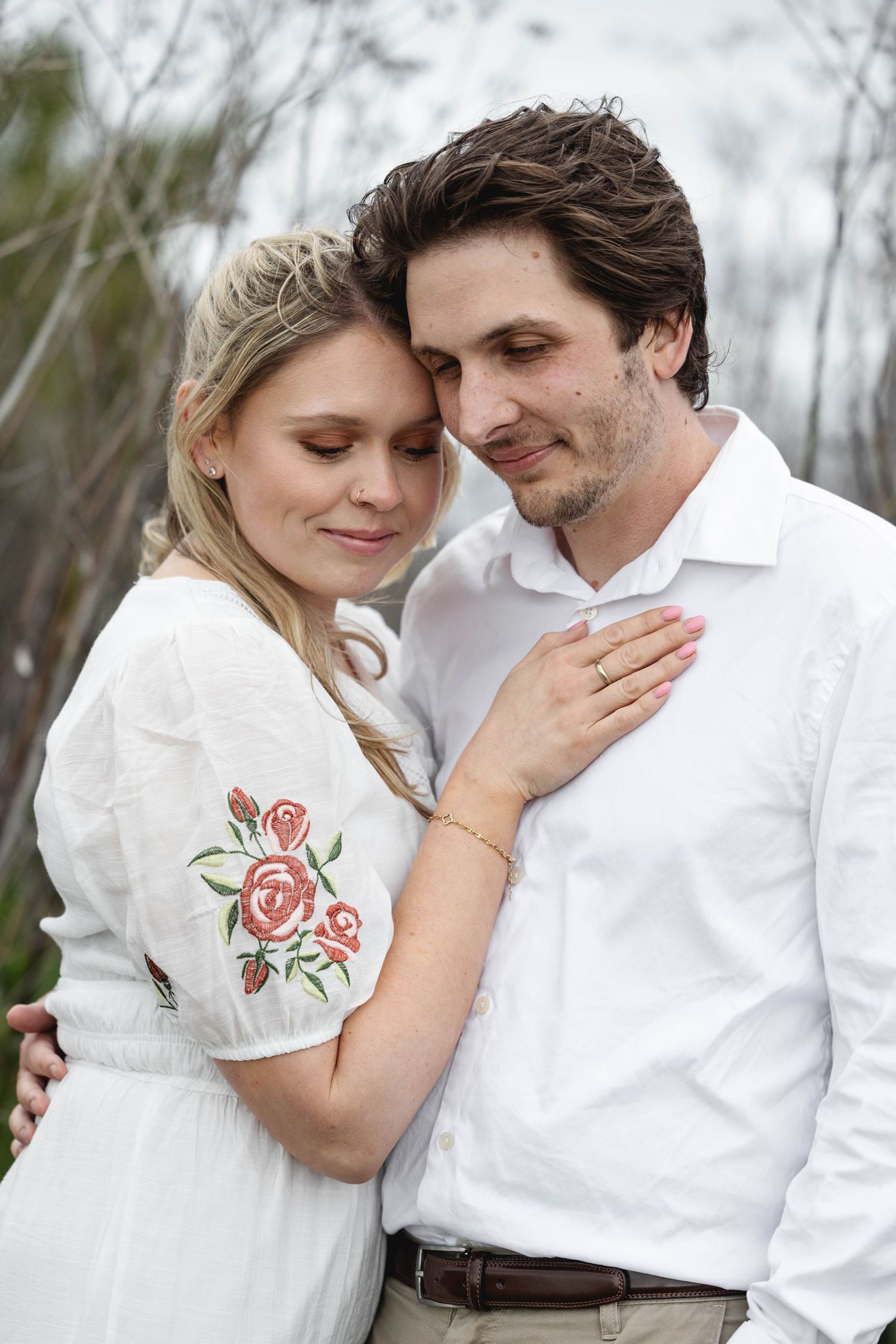 Romantic couple in white outfits embracing outdoors, woman in floral embroidered dress, man in white shirt, sharing tender moment.