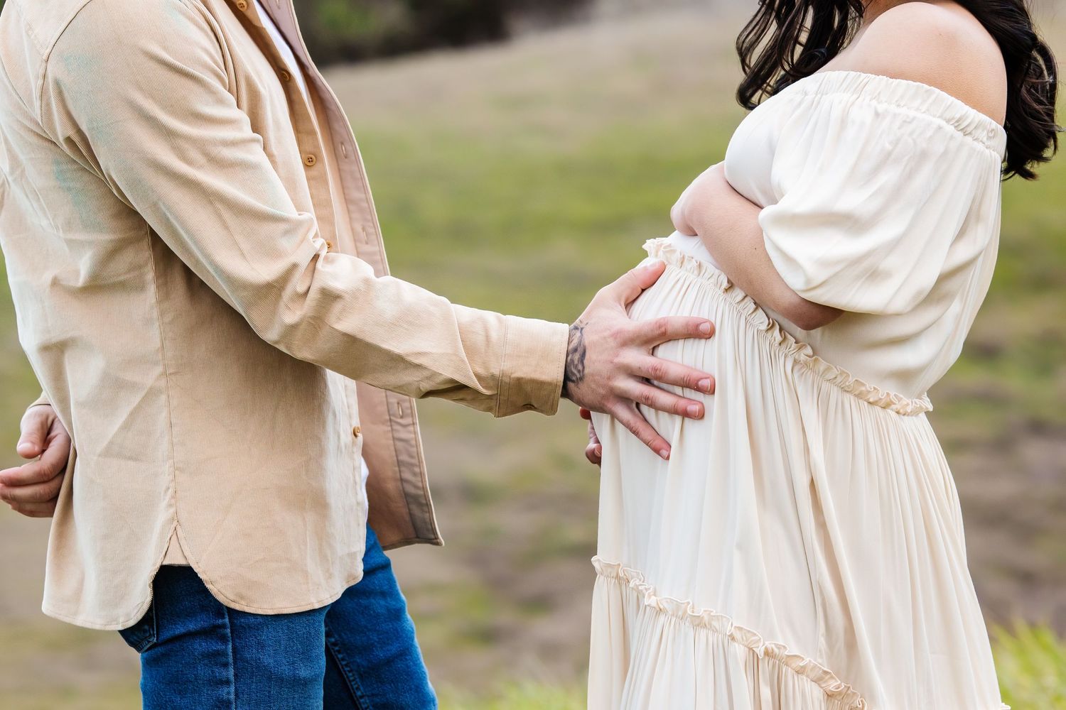 Couple sharing a tender maternity moment outdoors, man gently touching pregnant womans belly in neutral toned outfits.