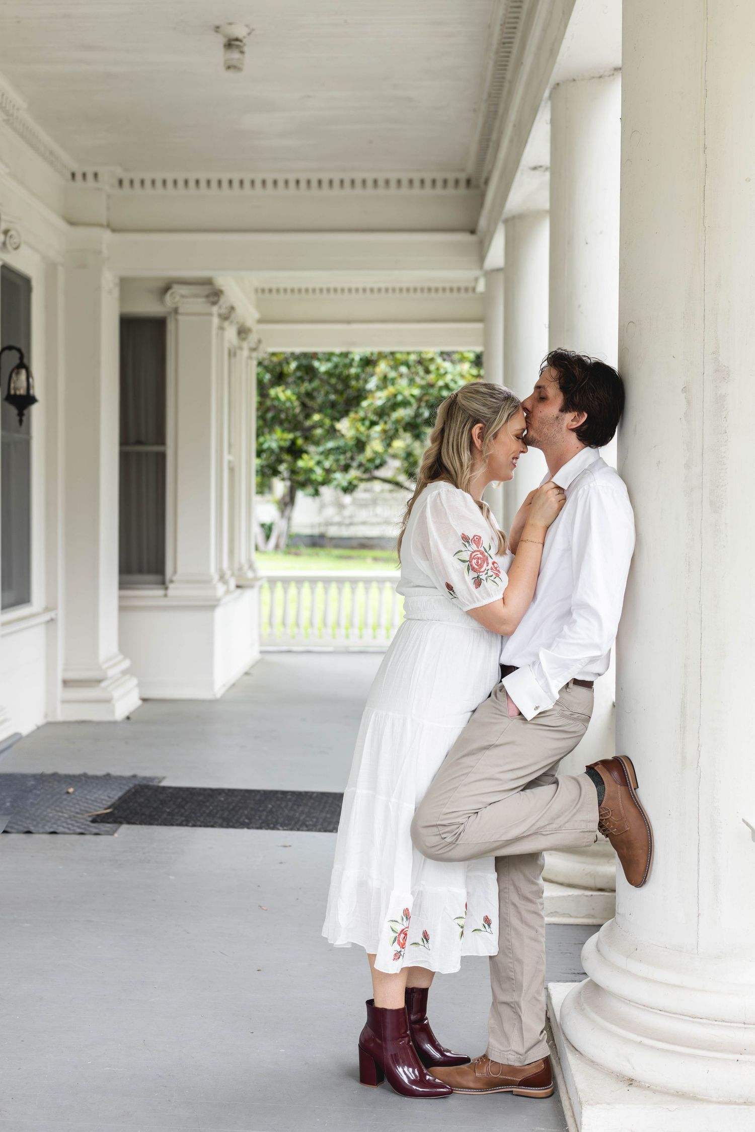 Romantic couple sharing an intimate moment on a white columned porch, woman in floral dress and man in white shirt.