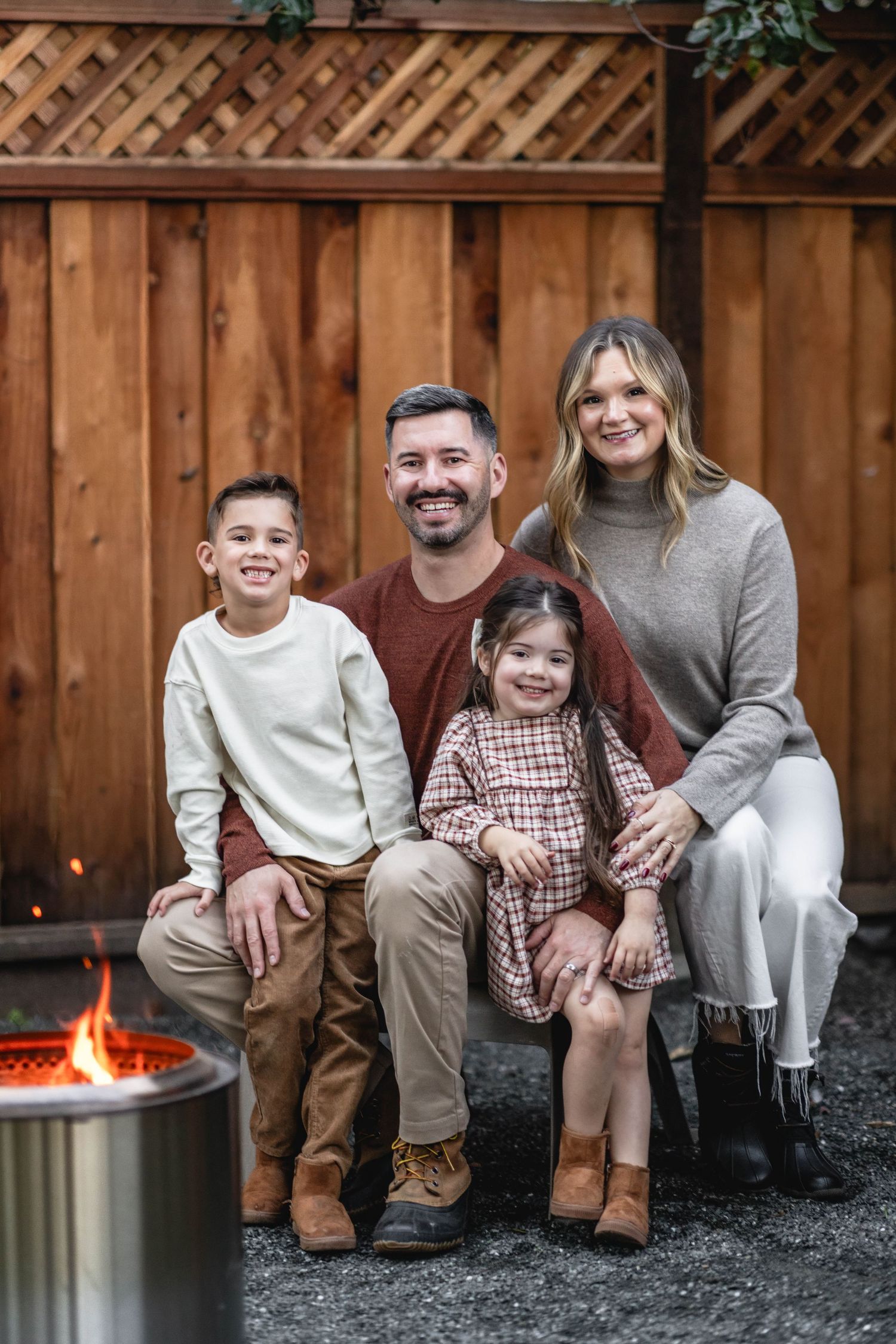 Family of four smiling together outdoors by a fire pit, with a wooden fence backdrop, dressed in warm autumn tones.