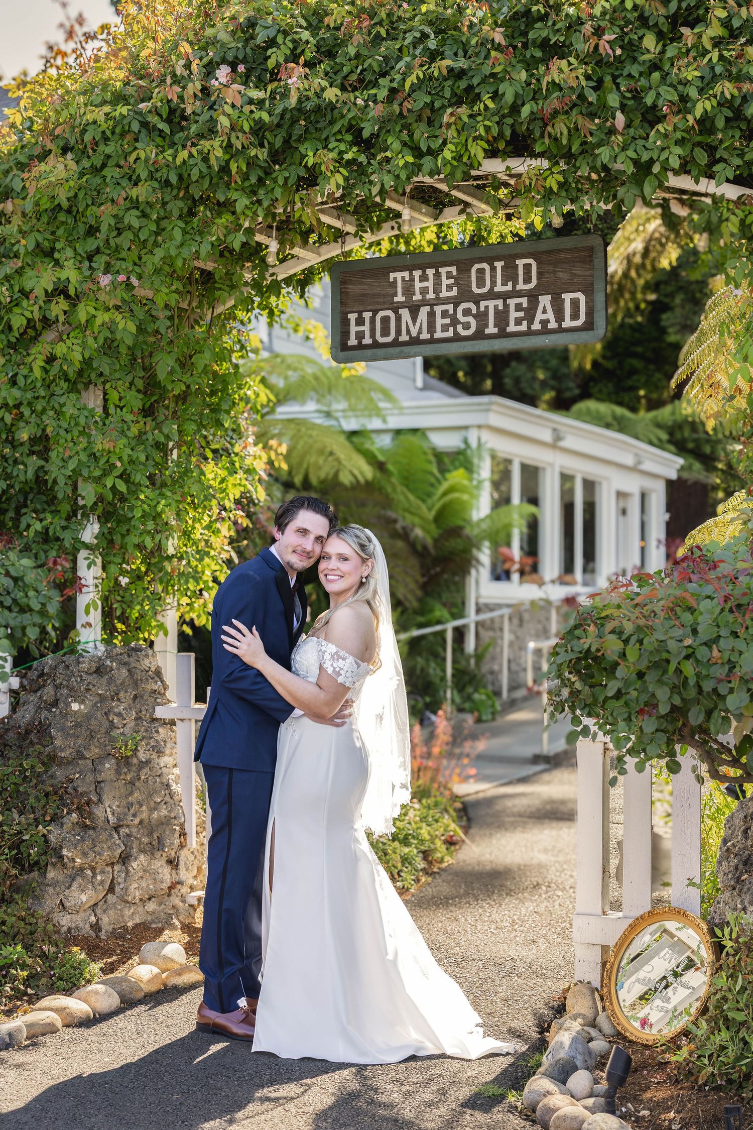 Bride and groom embrace under The Old Homestead sign, surrounded by lush greenery on their wedding day.