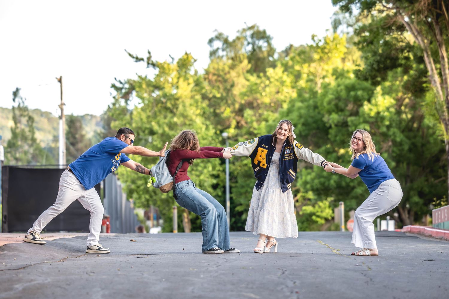 Four people playfully dancing and posing on an outdoor stage surrounded by lush green trees.