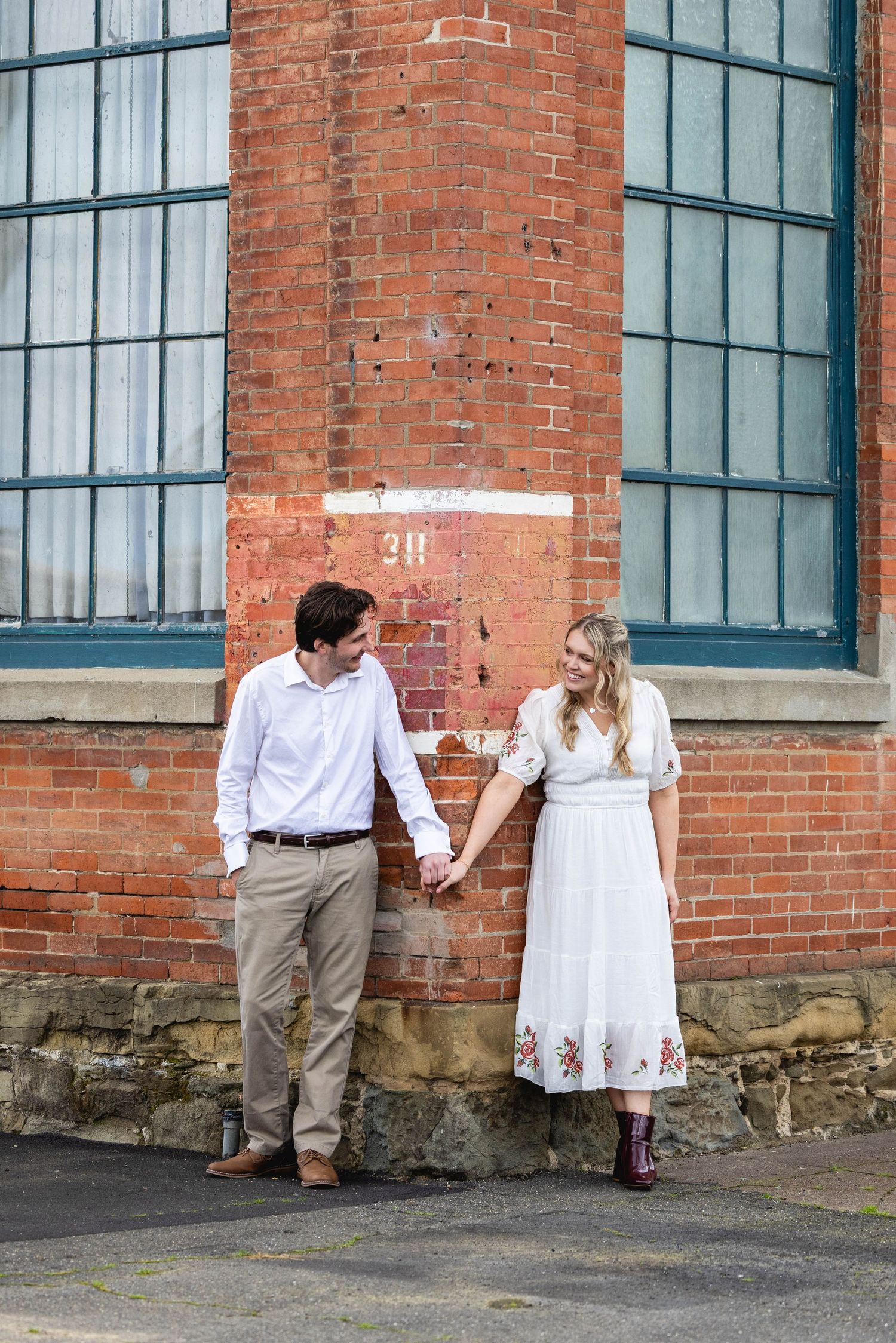 Couple holding hands in front of a red brick building, woman in white floral dress, man in white shirt and khaki pants.