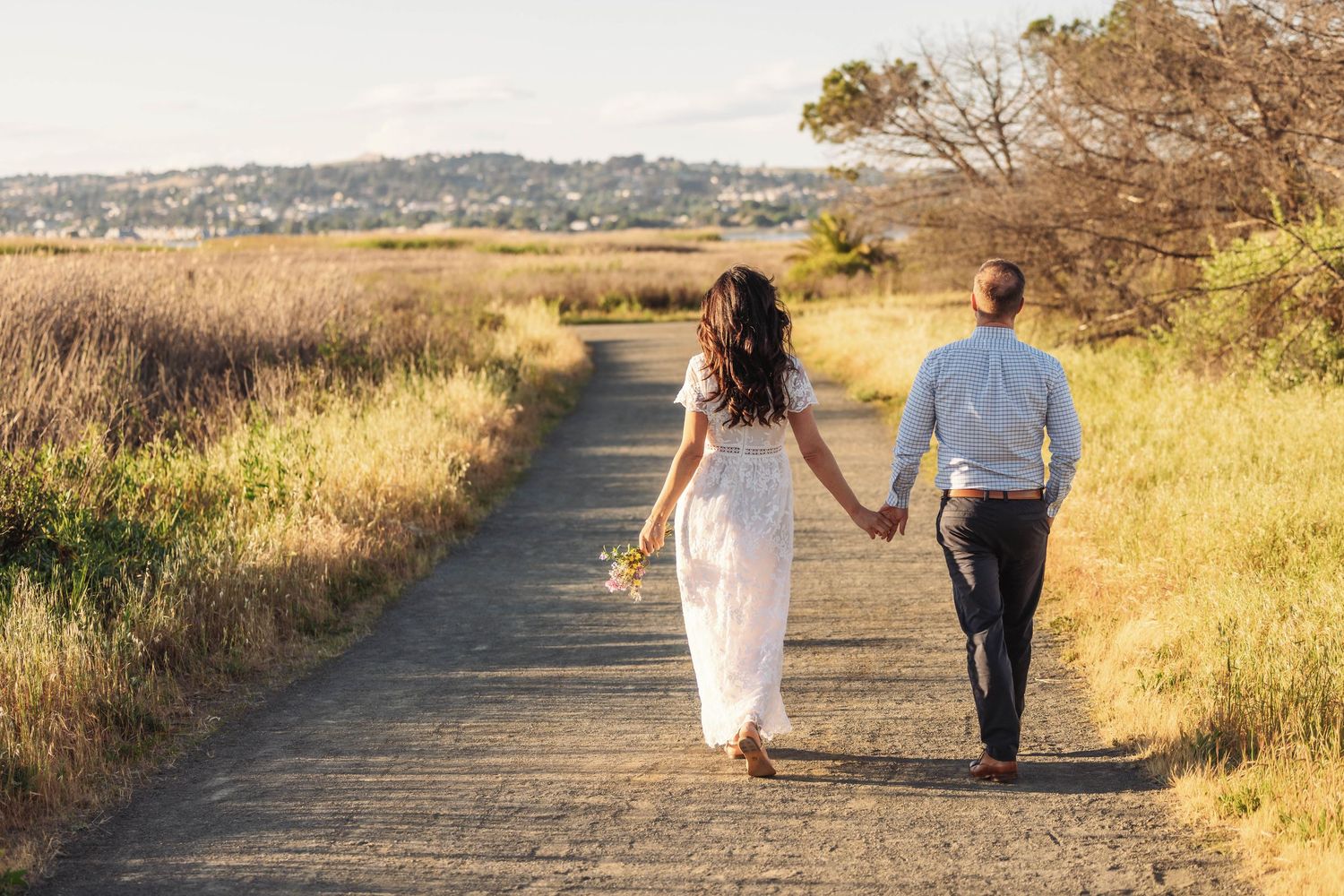 A couple in wedding attire walks hand-in-hand down a scenic path surrounded by golden fields.
