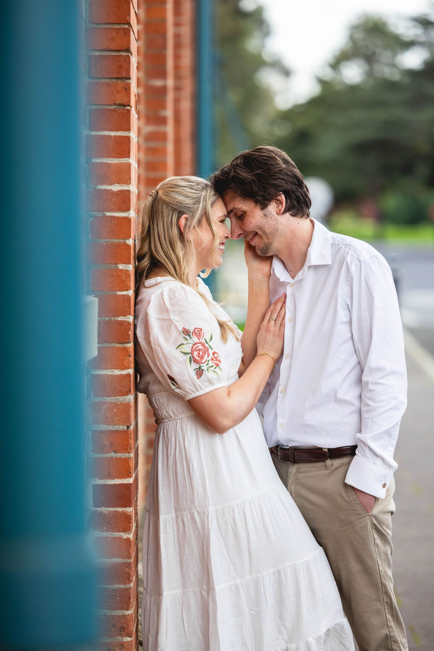 Couple sharing a romantic moment leaning against a brick wall, woman in white floral dress, man in white shirt.
