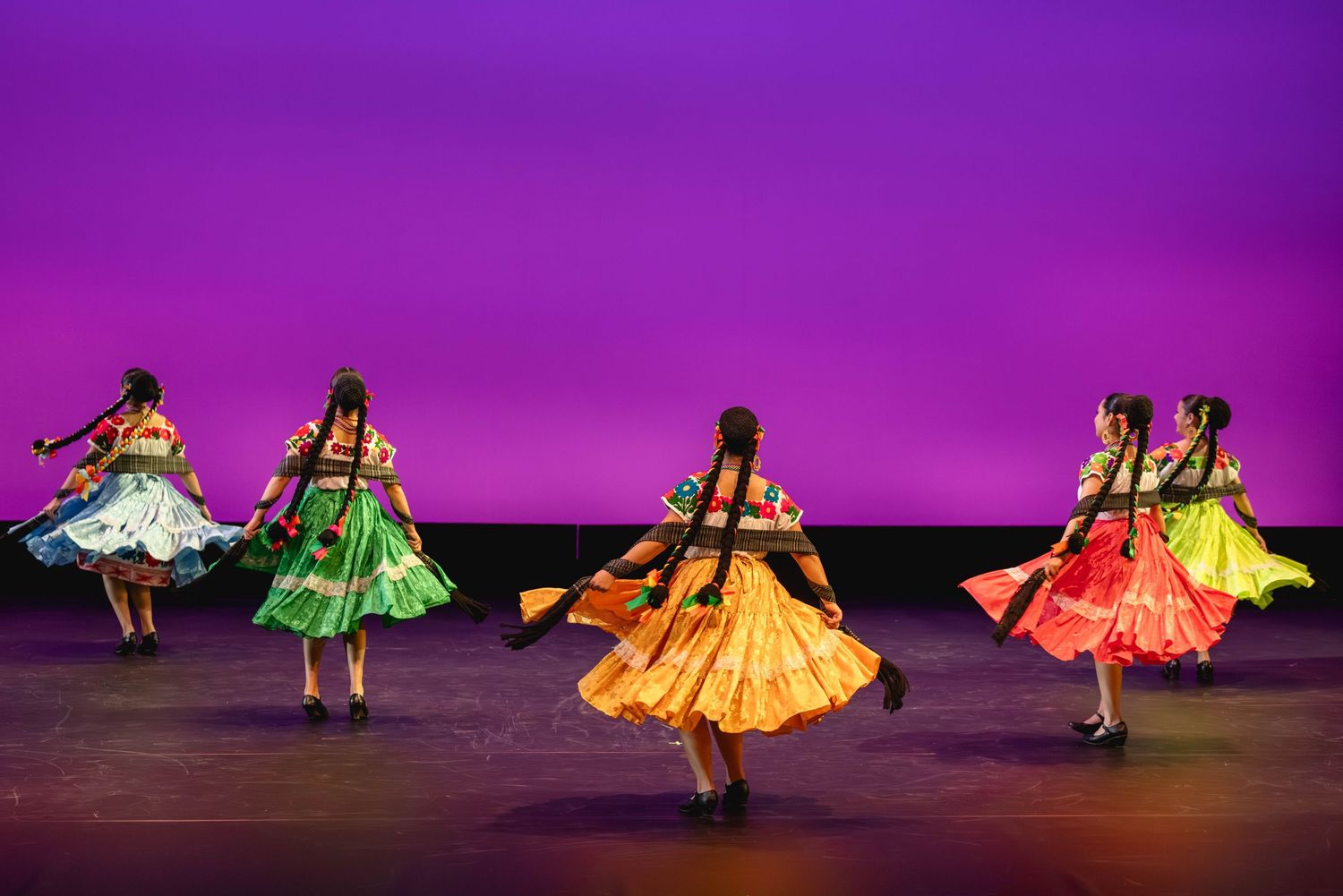 Colorful folkloric dancers in vibrant dresses perform on a purple-lit stage.