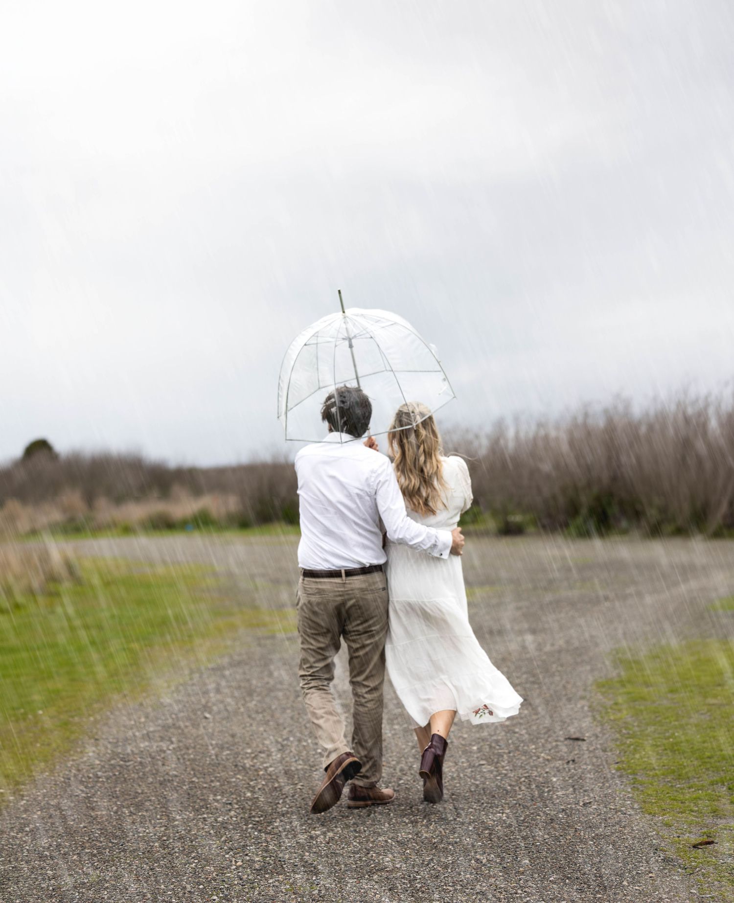 Couple walking in rain under clear umbrella on rural path, woman in white dress and man in khaki pants.