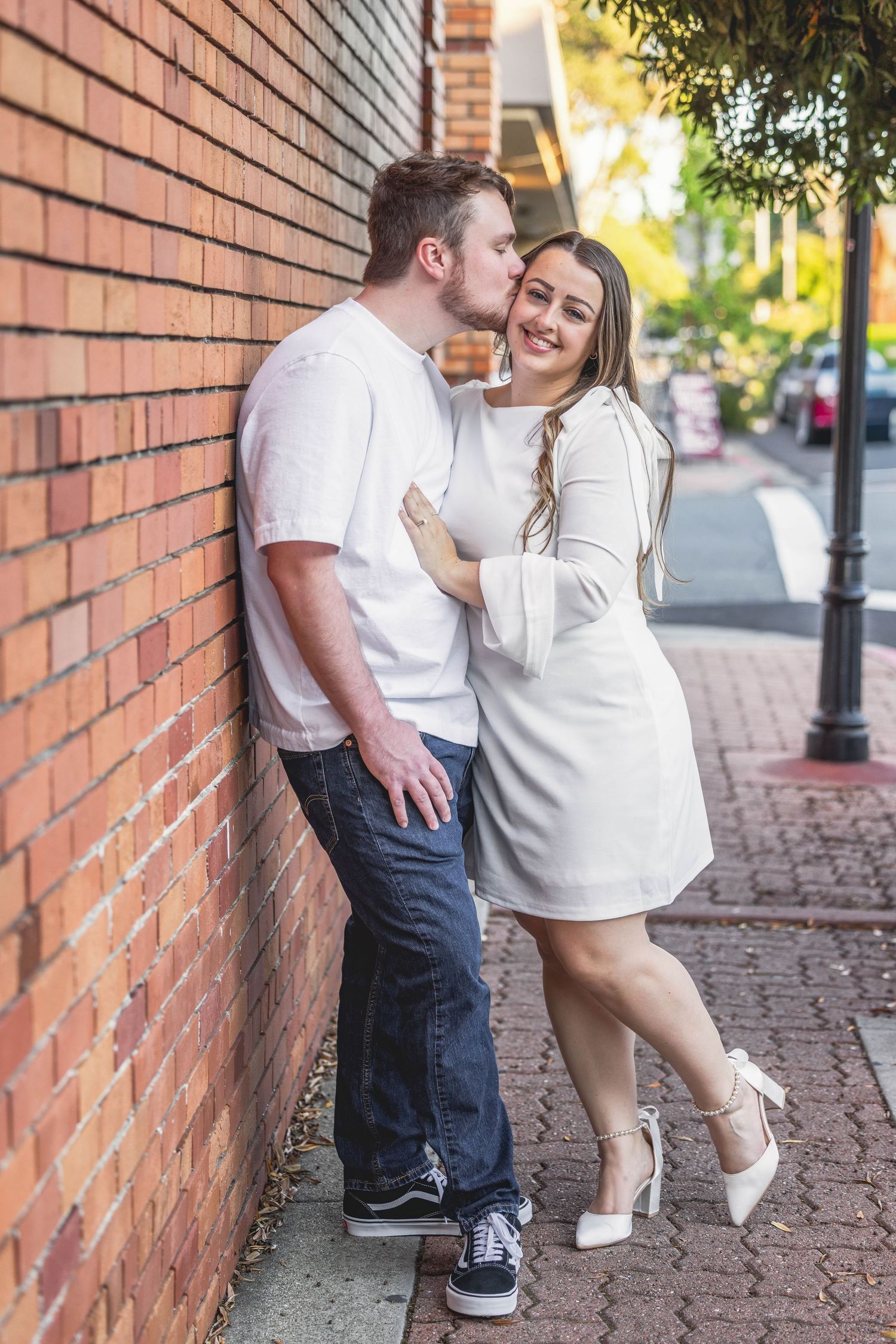 Couple in white outfits sharing a romantic kiss against a brick wall on a sunny urban sidewalk.