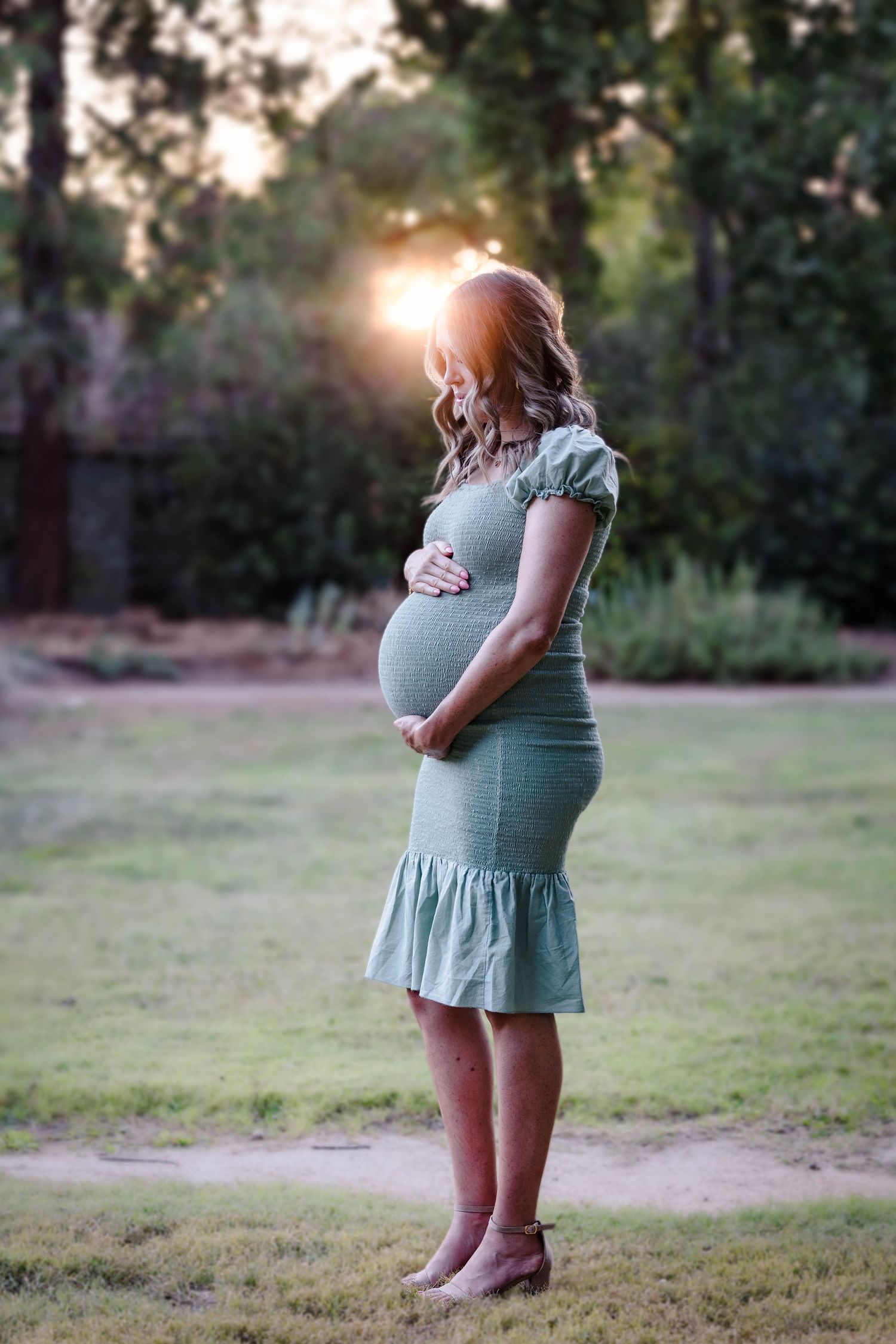Pregnant woman in sage green dress cradling baby bump outdoors at golden hour sunset in lush garden setting.