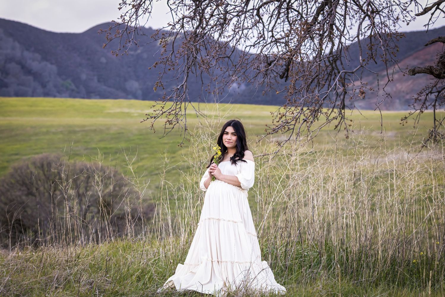 Woman in white dress holding flower in green meadow with mountains and bare tree branches in background.