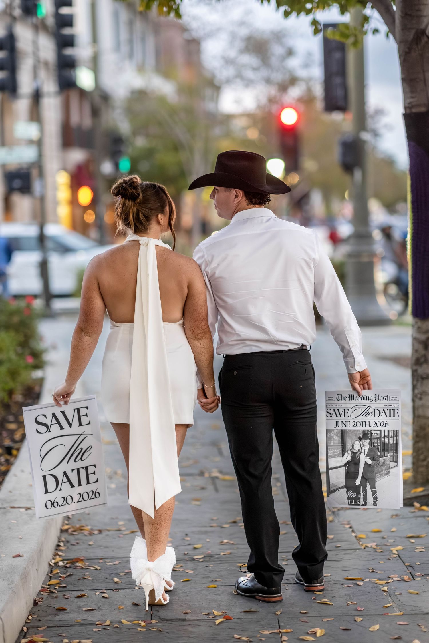 Couple walking hand-in-hand on sidewalk holding Save the Date signs, bride in white dress, groom in cowboy hat.