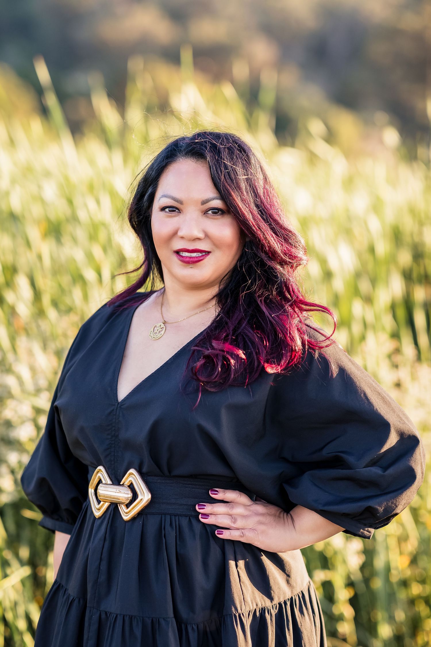 A woman in a black dress poses outdoors in golden sunlight against a natural backdrop of tall grass.