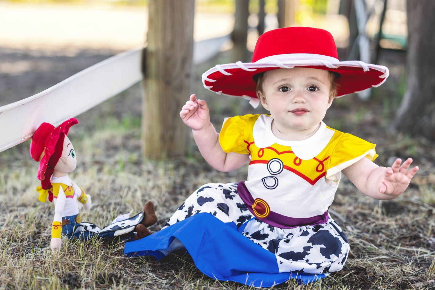 Baby dressed as Jessie from Toy Story sitting outdoors next to a Jessie doll, wearing red hat and cowgirl outfit.