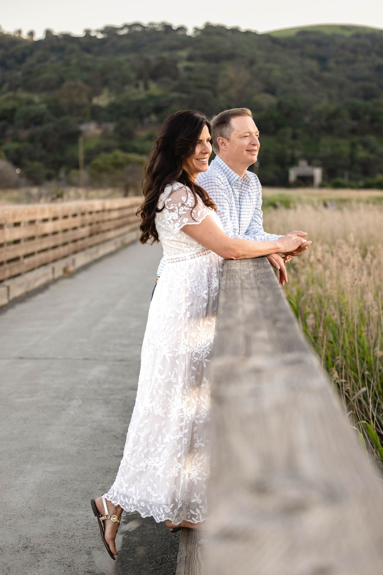 Couple in white lace dress and blue shirt leaning on wooden bridge railing with marsh grass and green hills behind.