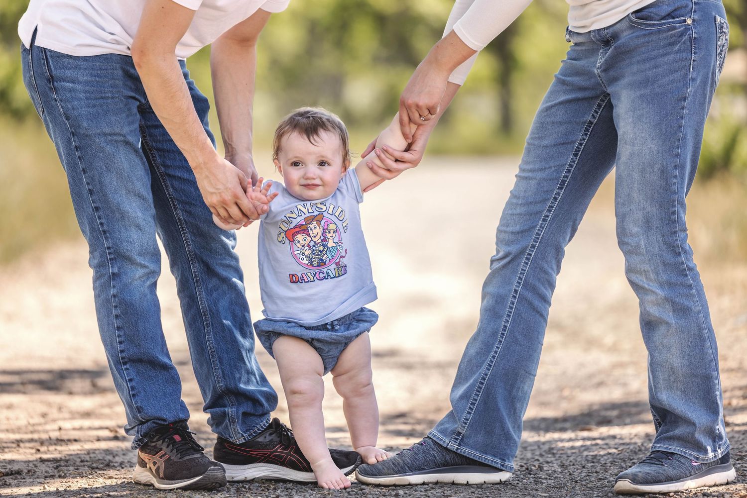 Two parents holding hands of a baby learning to walk outdoors on a gravel path in summer.