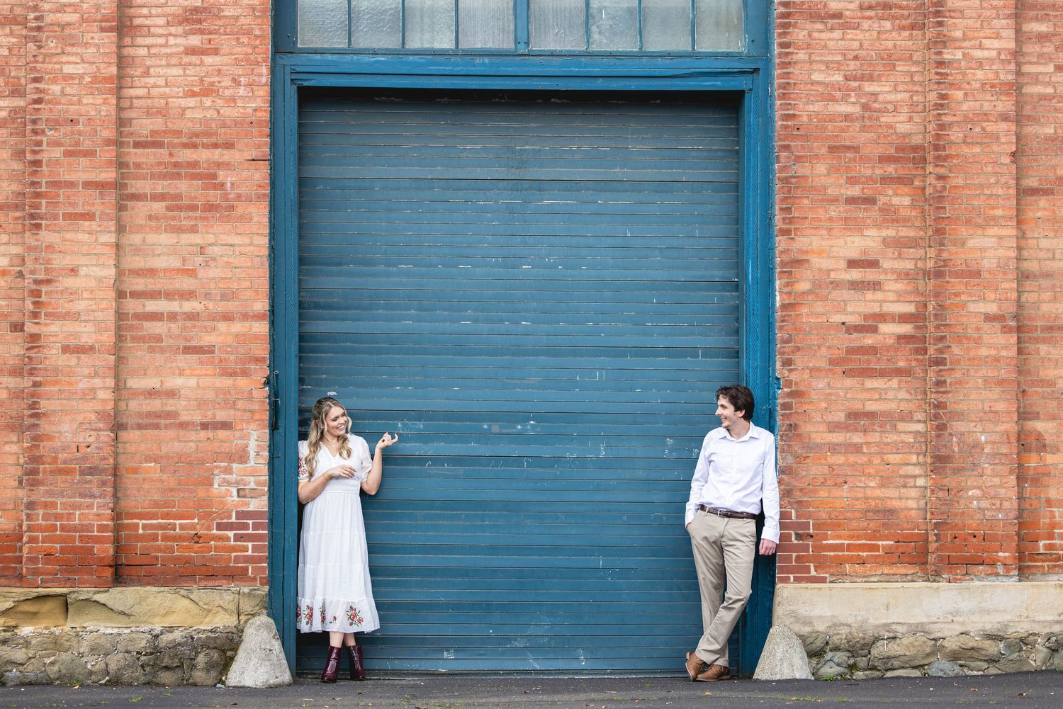 Couple standing apart by a blue garage door on a brick building, woman in white dress, man in white shirt.