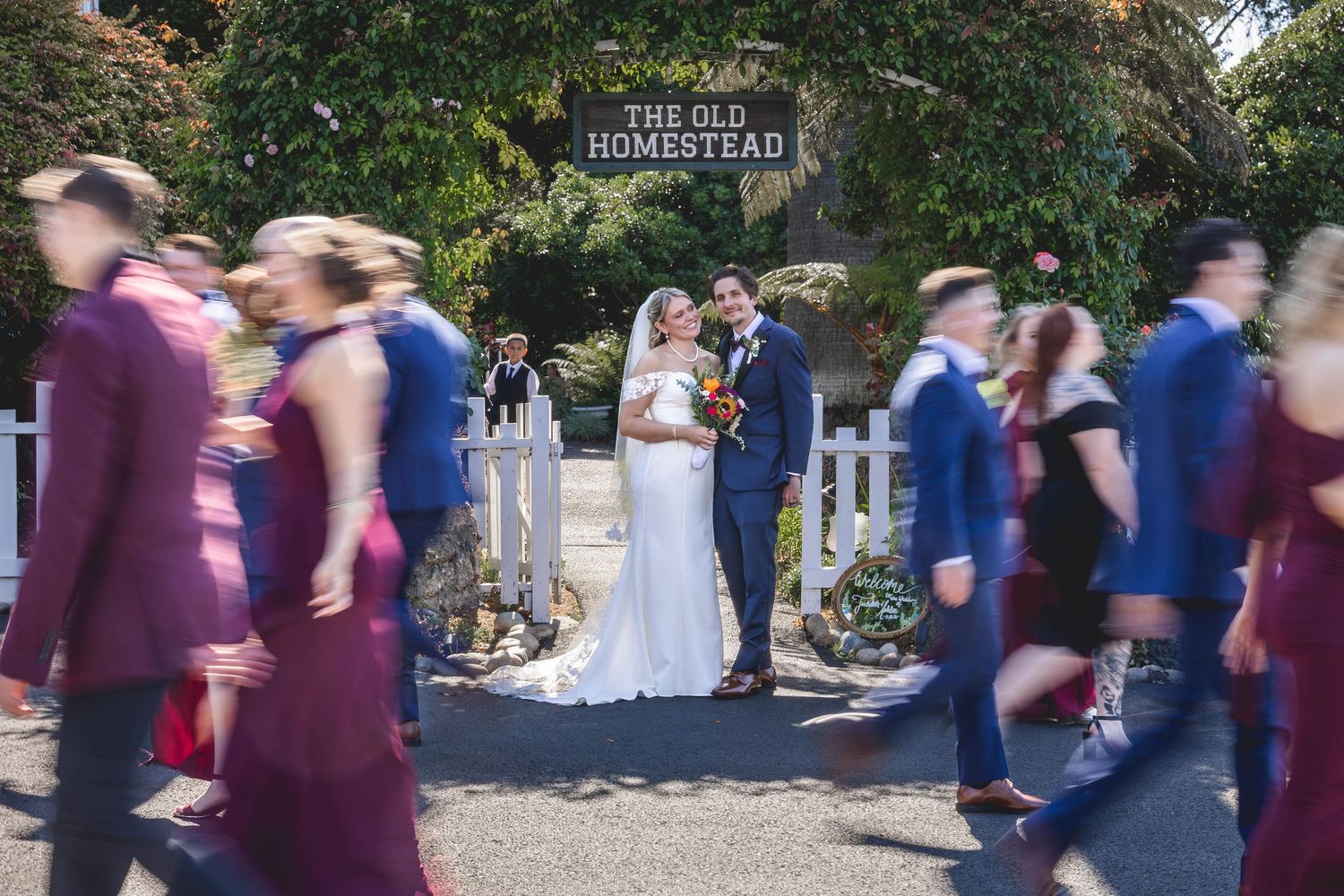 Bride and groom pose at The Old Homestead venue entrance while wedding party walks past in blue suits and burgundy dresses.