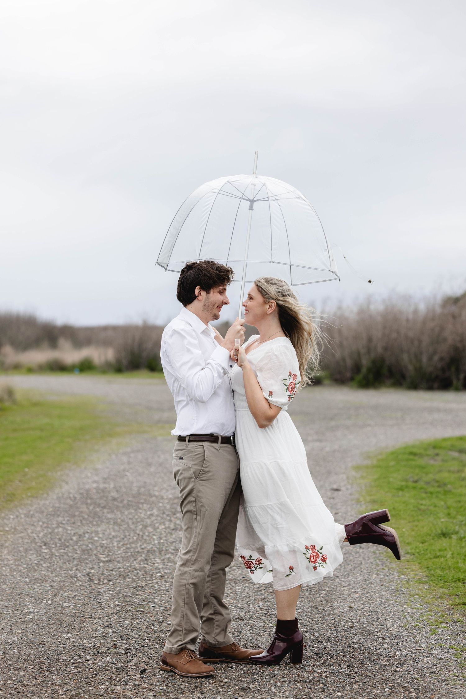 Couple sharing a romantic moment under a clear umbrella on a gravel path, woman in white floral dress, man in white shirt.