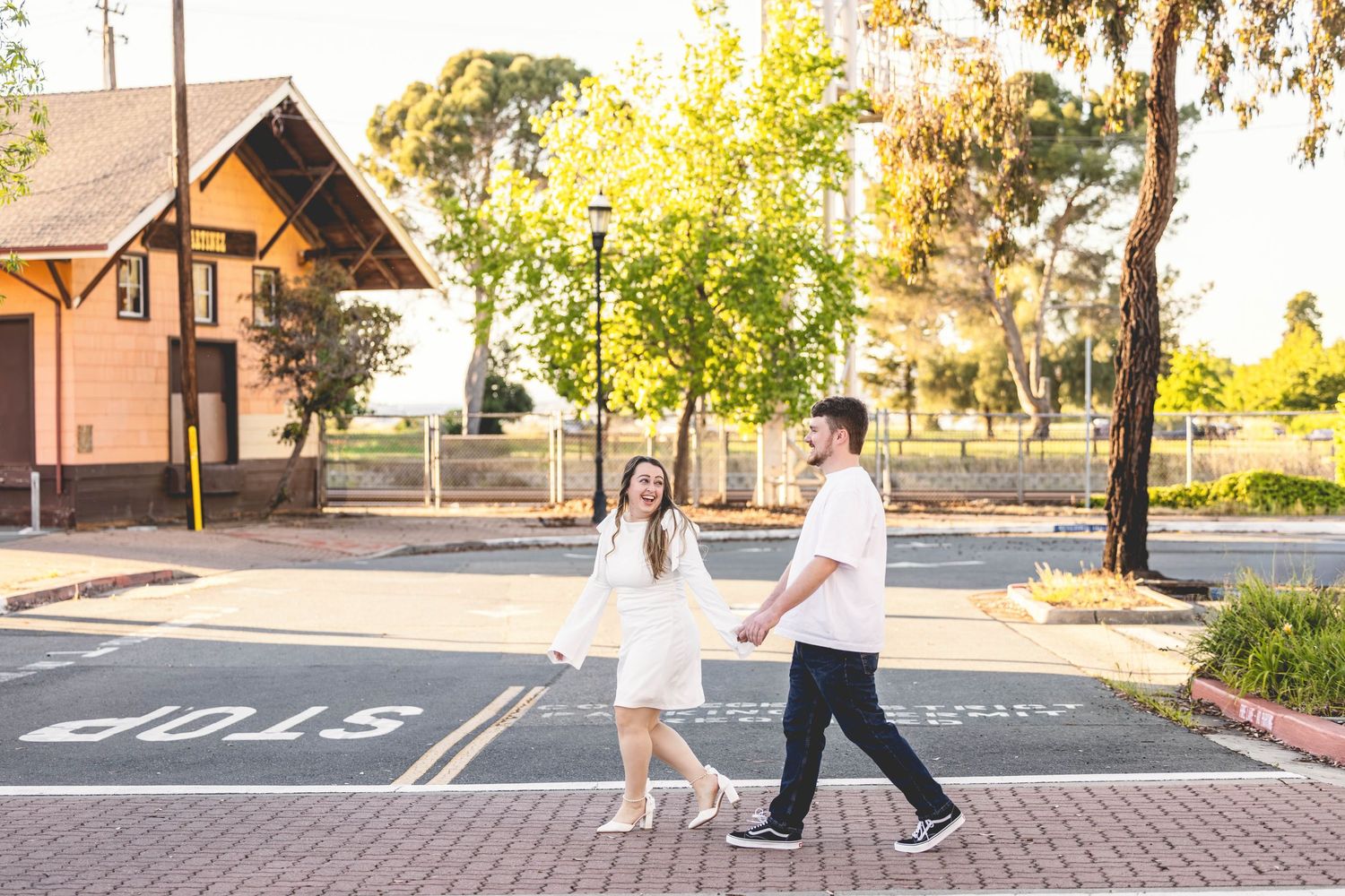 Couple holding hands crossing a street at golden hour near a historic train station building.