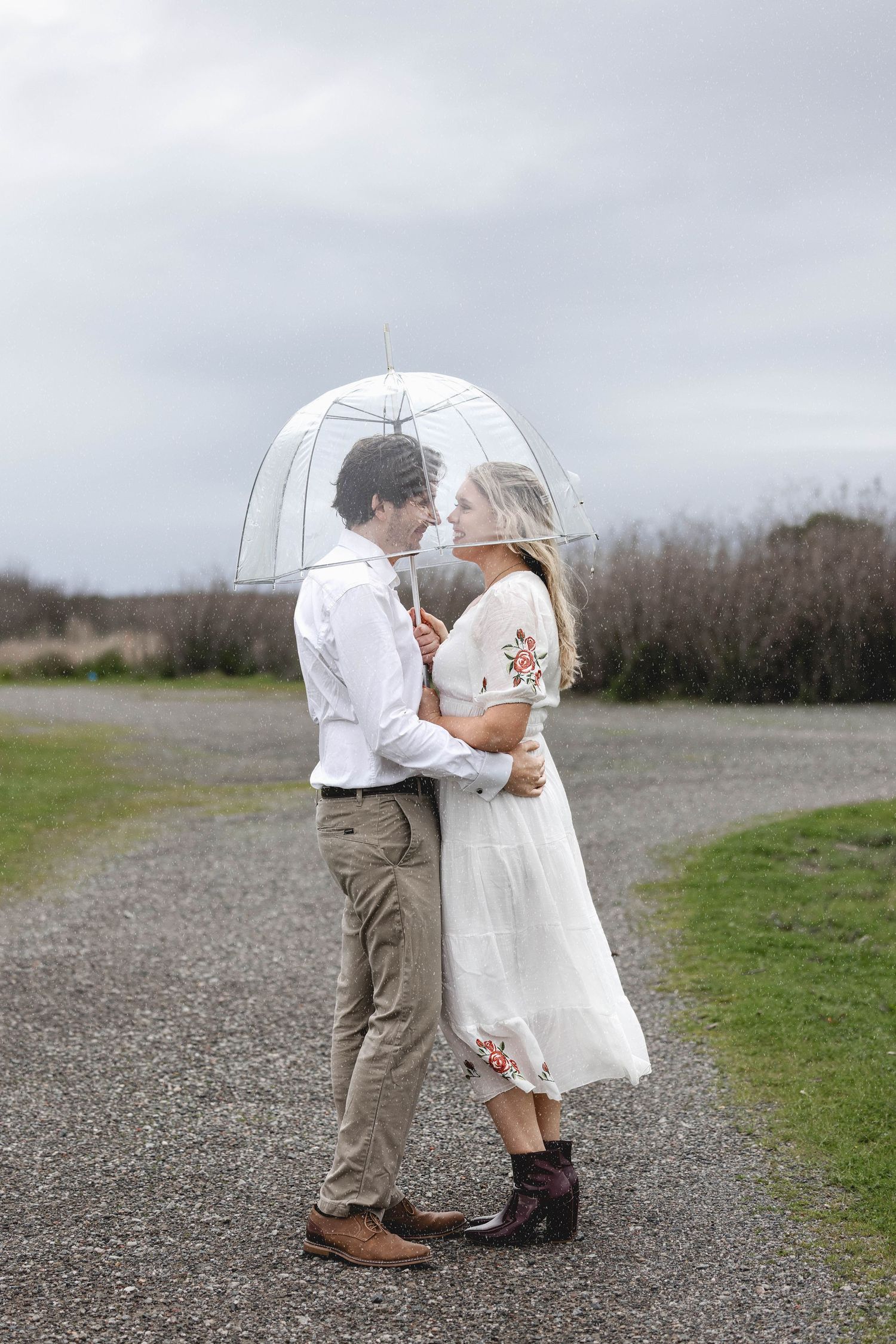 Couple sharing a romantic moment under a clear bubble umbrella on a gravel path on a cloudy day.