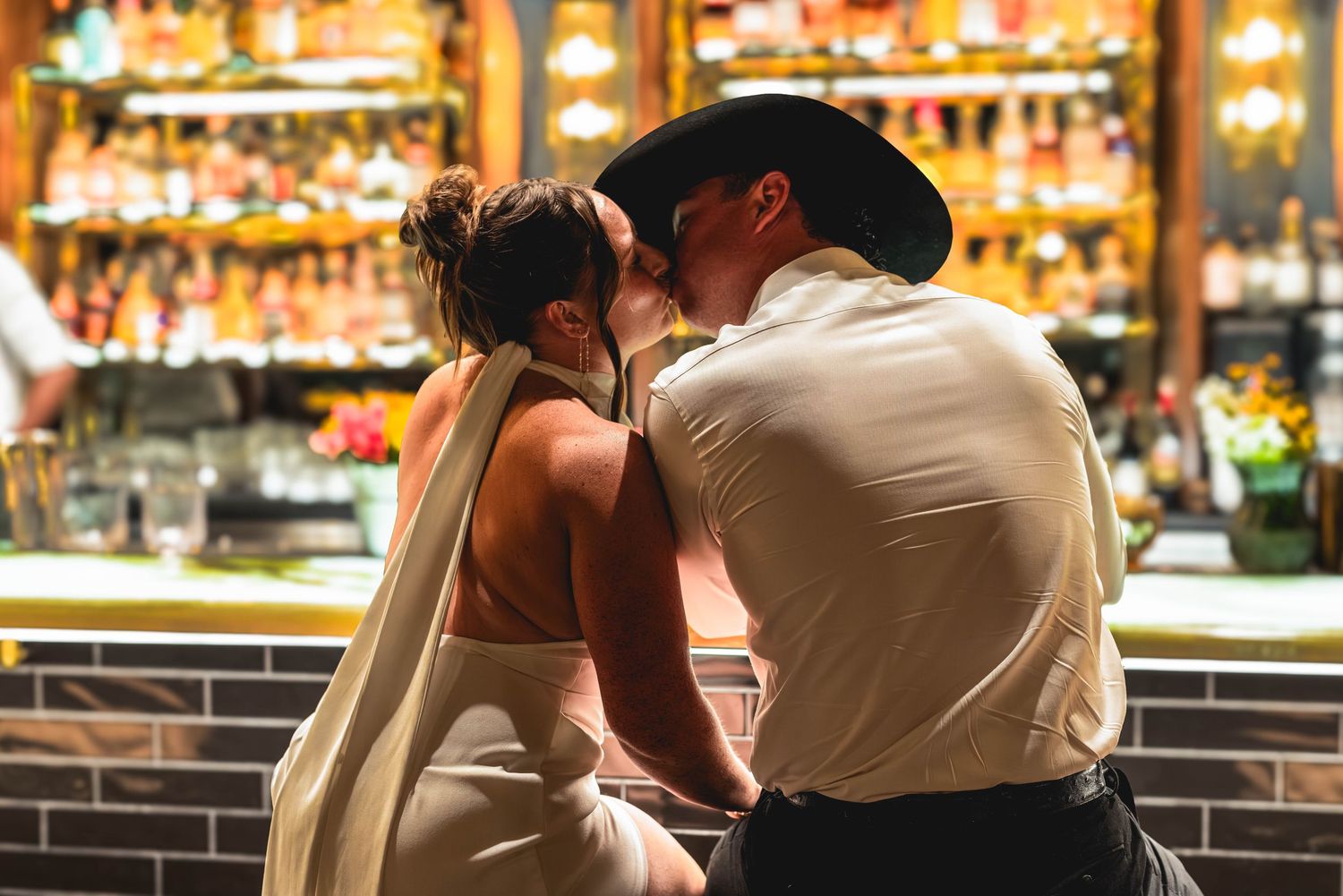 Couple sharing a romantic kiss at a warmly lit bar, woman in backless dress, man in cowboy hat.