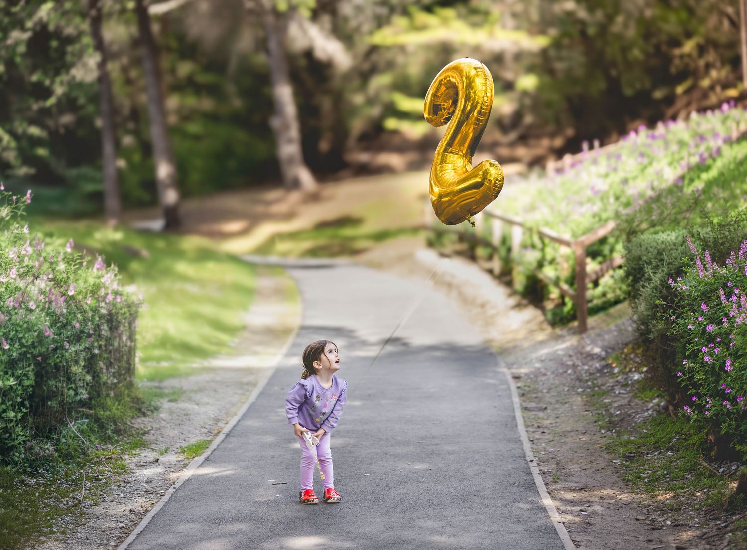 A toddler stands on a park path holding a gold number 2 balloon among lush green trees.
