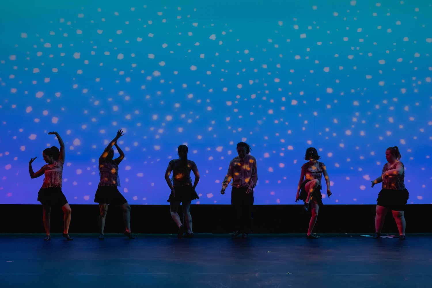Six dancers silhouetted against a blue bokeh backdrop performing on a dimly lit stage.