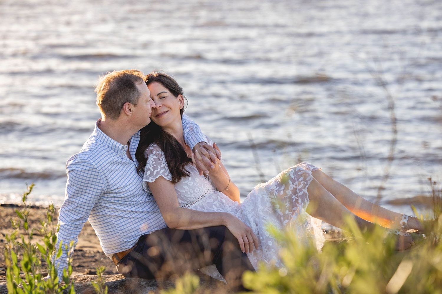 Couple sitting together on a sandy riverbank, sharing a romantic moment in warm golden sunlight.