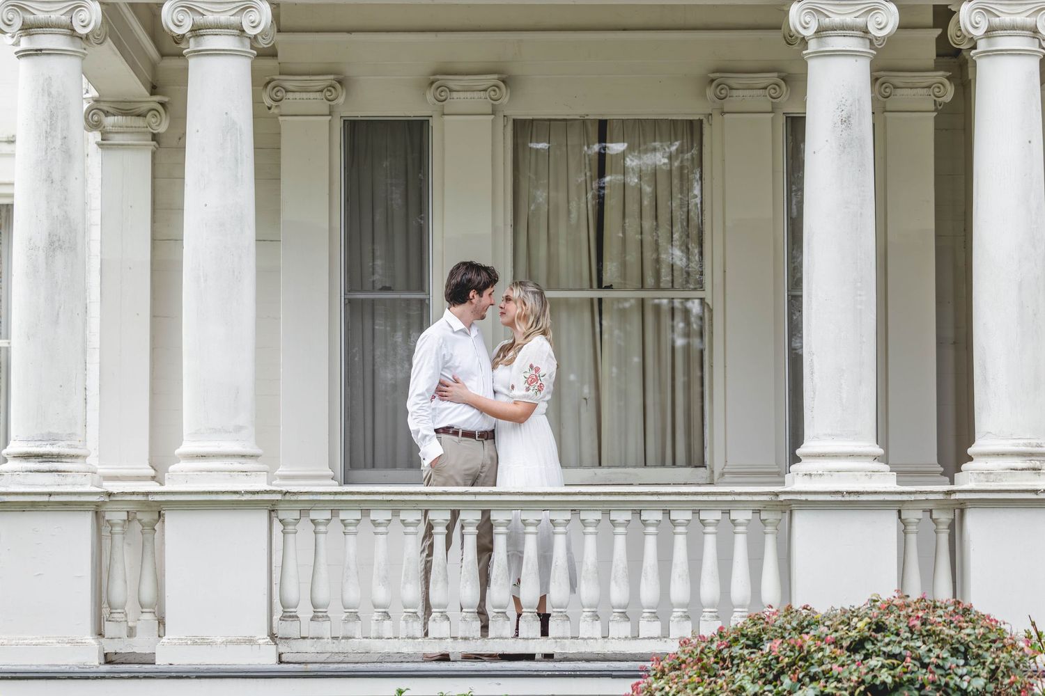 Couple sharing a romantic moment on a white columned porch of an elegant Southern mansion.