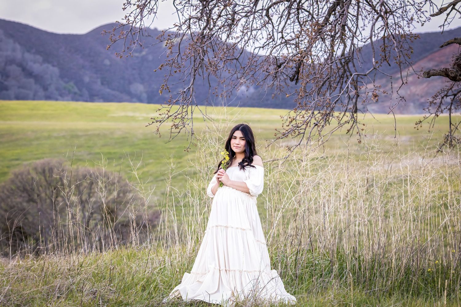 Woman in white dress holding flower in green meadow with mountains and bare tree branches in background.