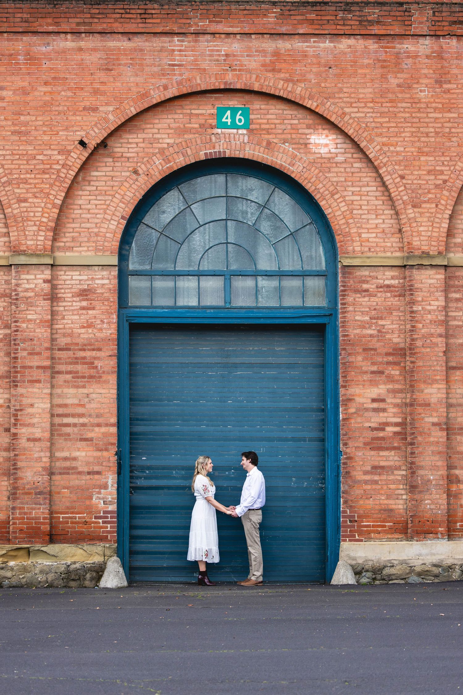Couple holding hands in front of large teal door numbered 46 on a red brick building.