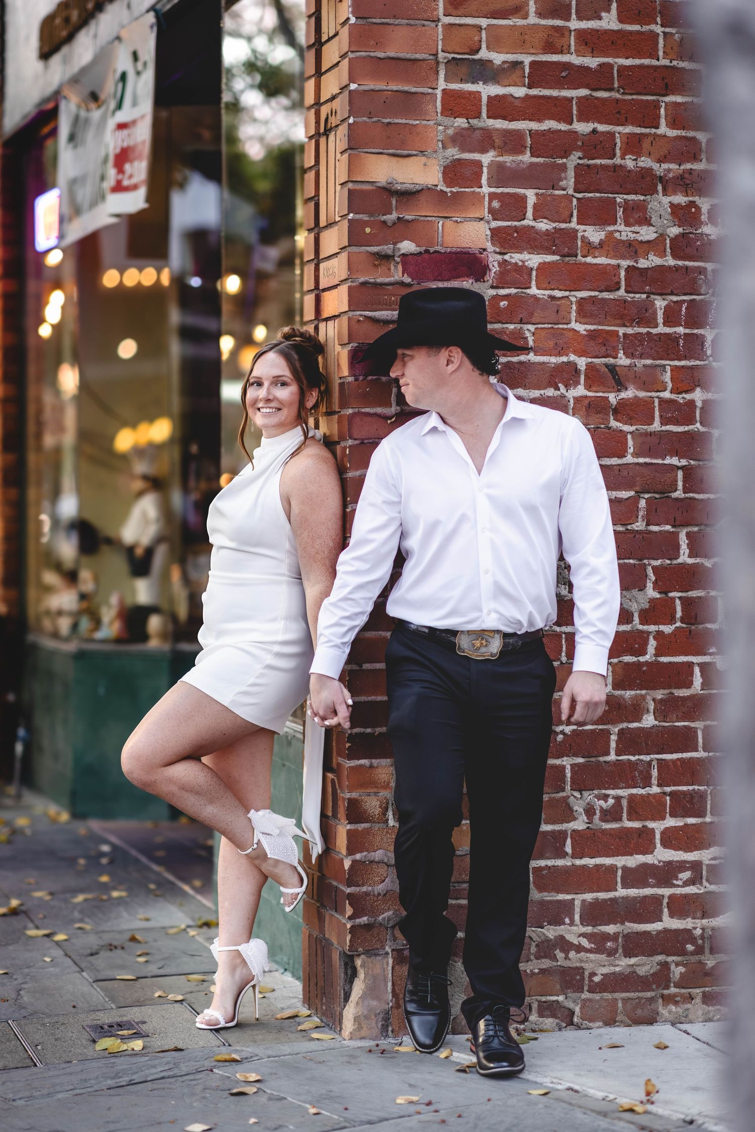 Couple in white dress and cowboy hat leaning against brick wall on urban sidewalk.