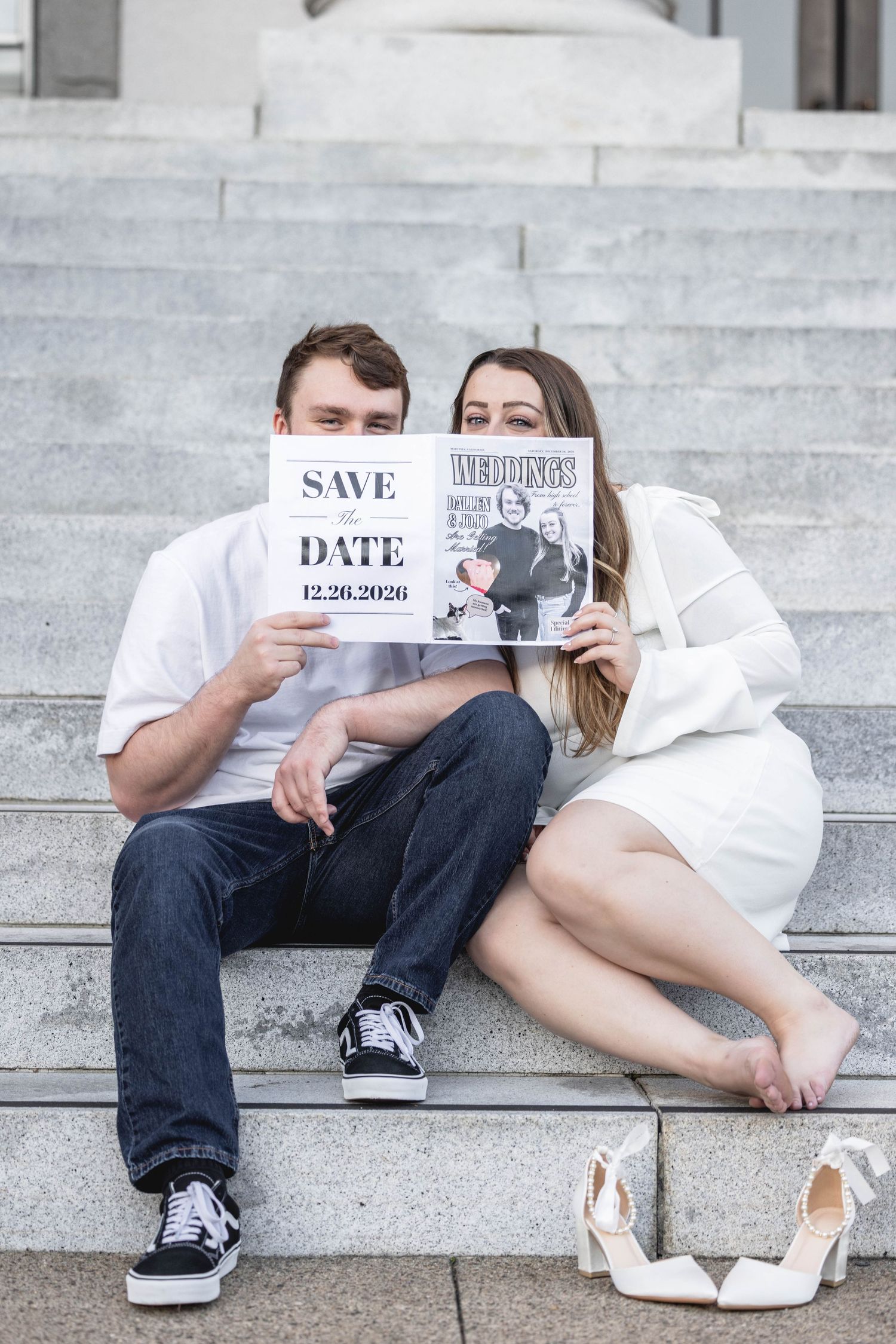 Couple sitting on stone steps holding Save the Date card and wedding magazine, dressed in white and jeans.
