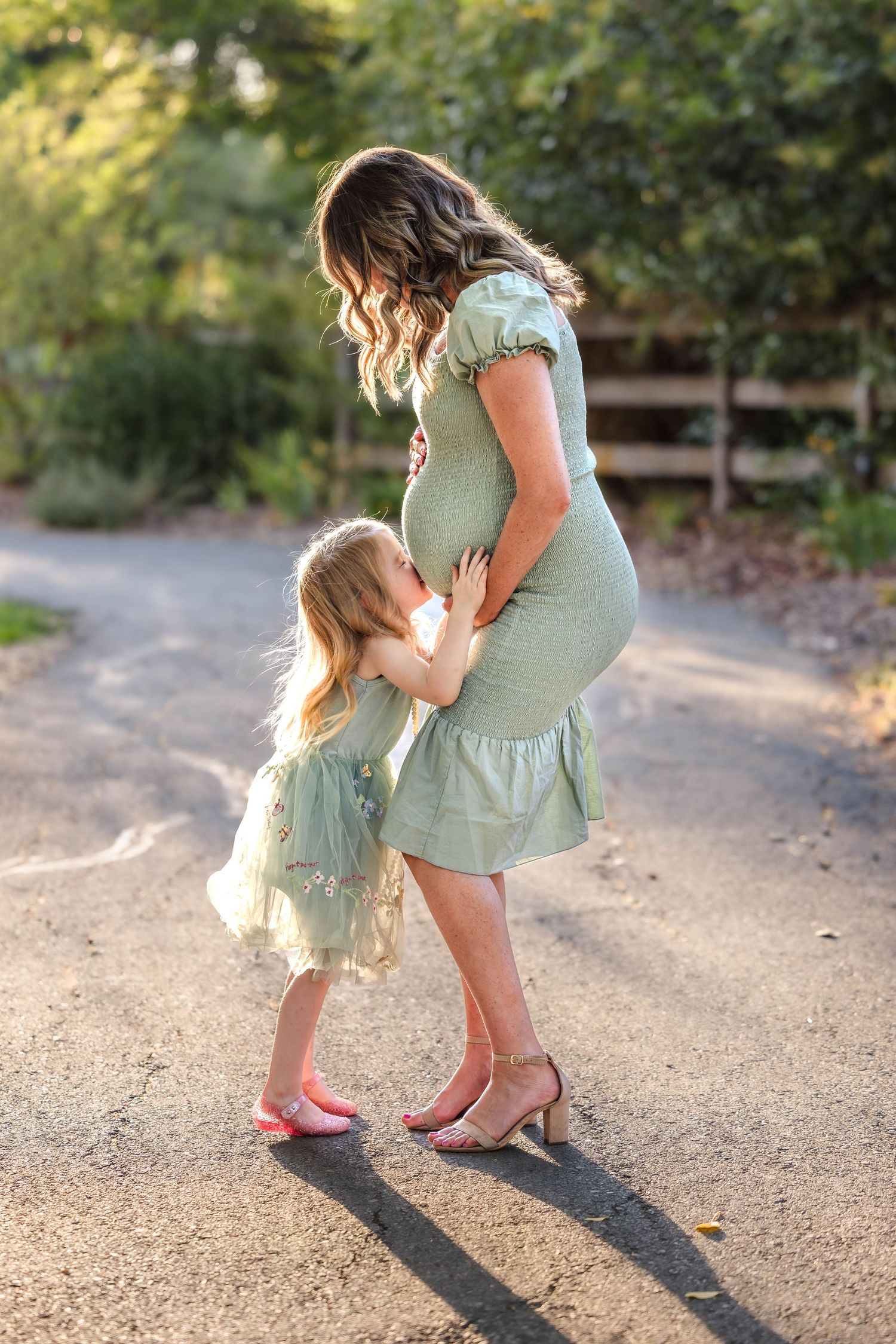 Pregnant woman in sage green dress with young daughter hugging her baby bump outdoors on a sunlit path.