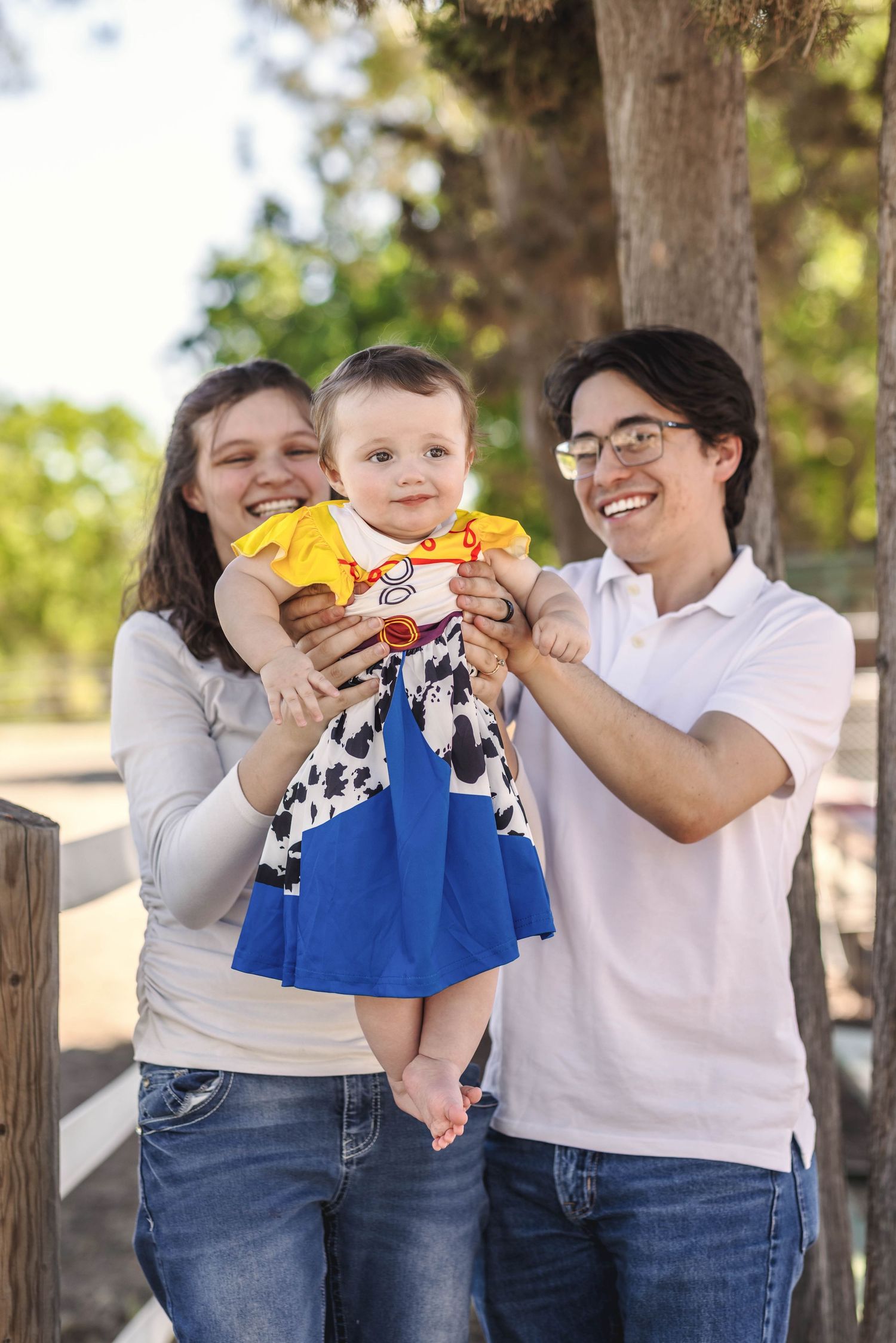 Happy young family with parents holding a baby dressed in a Toy Story Jessie costume outdoors near a wooden fence.