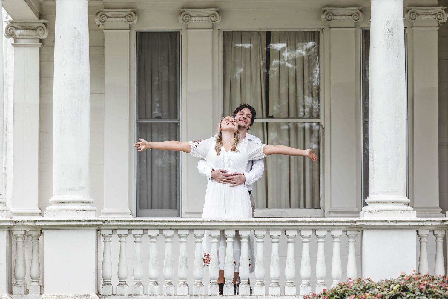 Joyful couple embracing on a grand white columned veranda, woman spreading her arms wide.