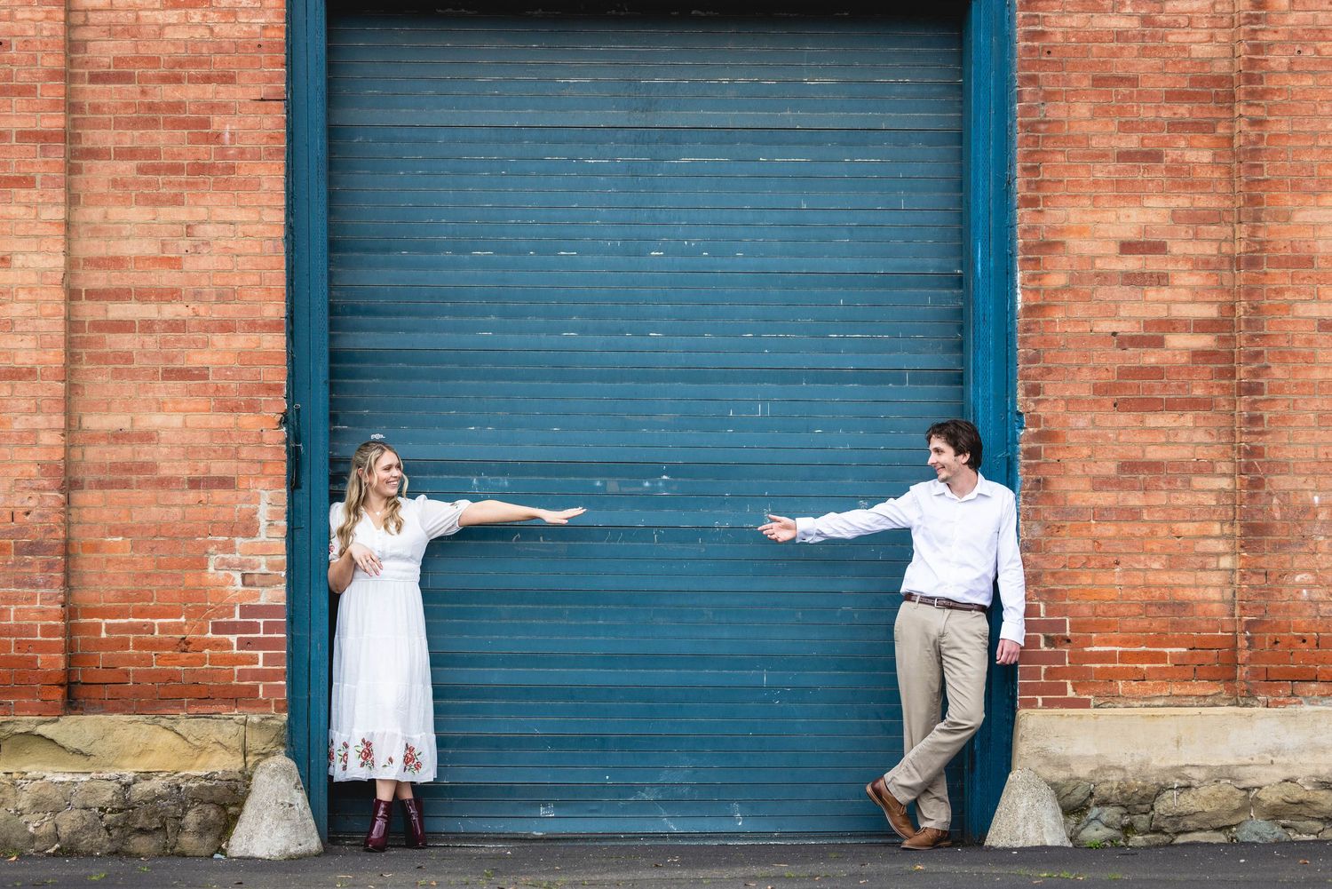 Couple in white outfits reaching toward each other in front of a teal garage door on a brick wall.