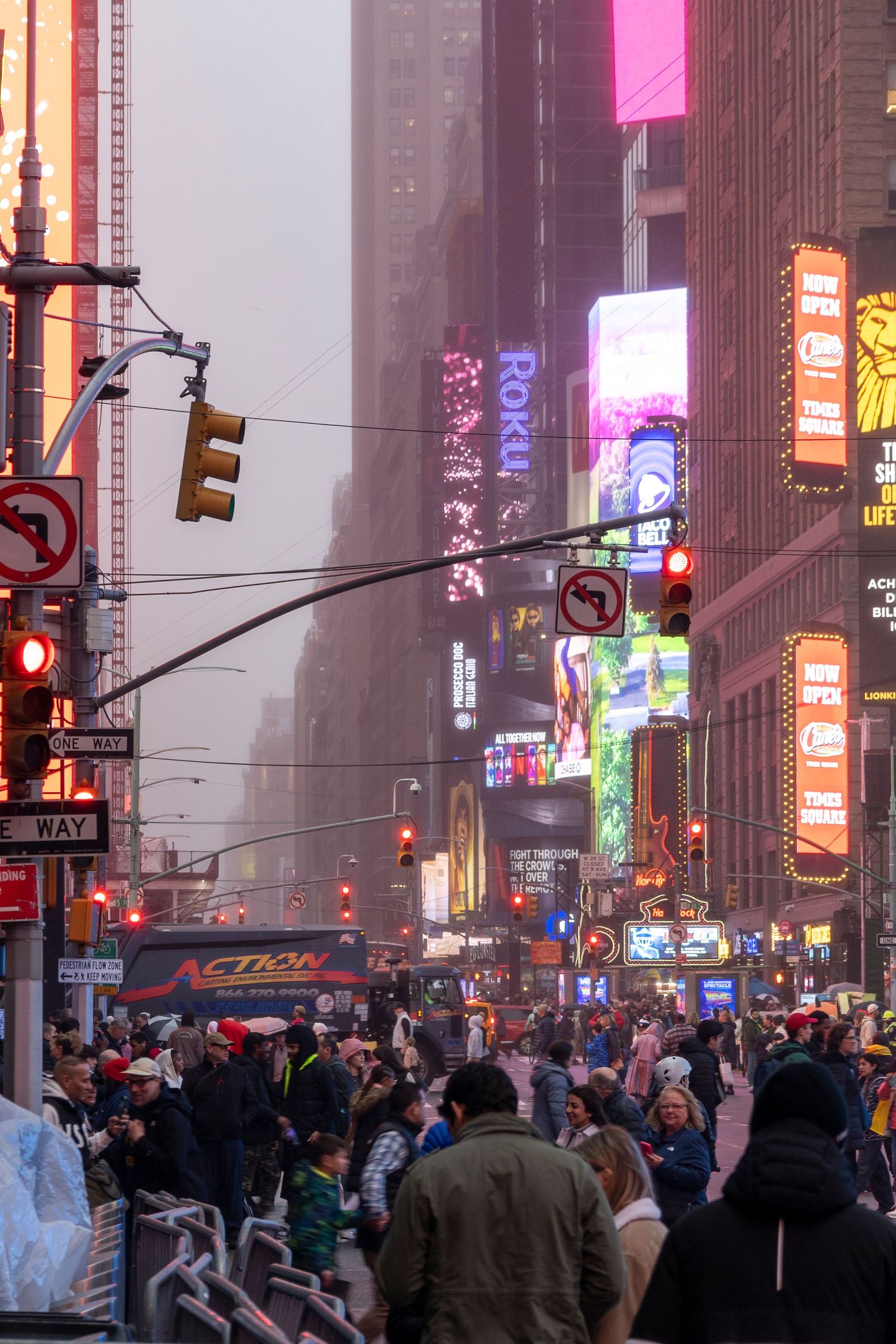 A bustling crowd walks through Times Square at dusk with bright neon signs and billboards illuminating the foggy NYC street.