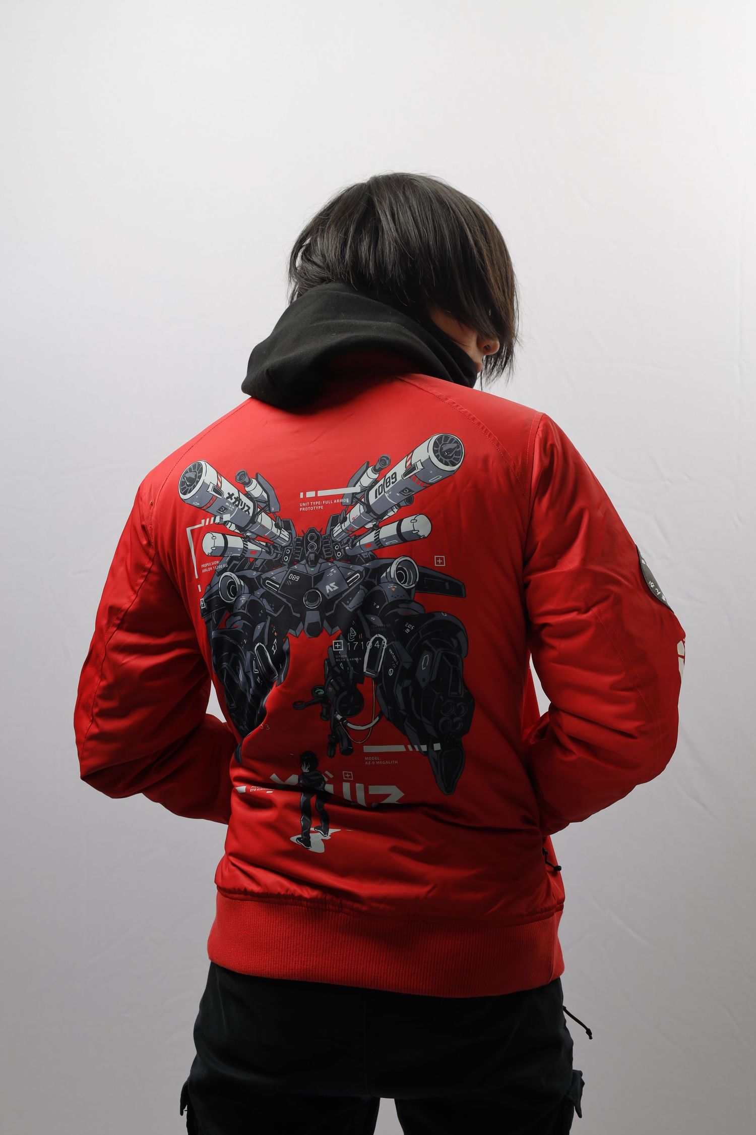 A person wearing a red sweater with a robot design on the back and a black cap stands against a gray background.