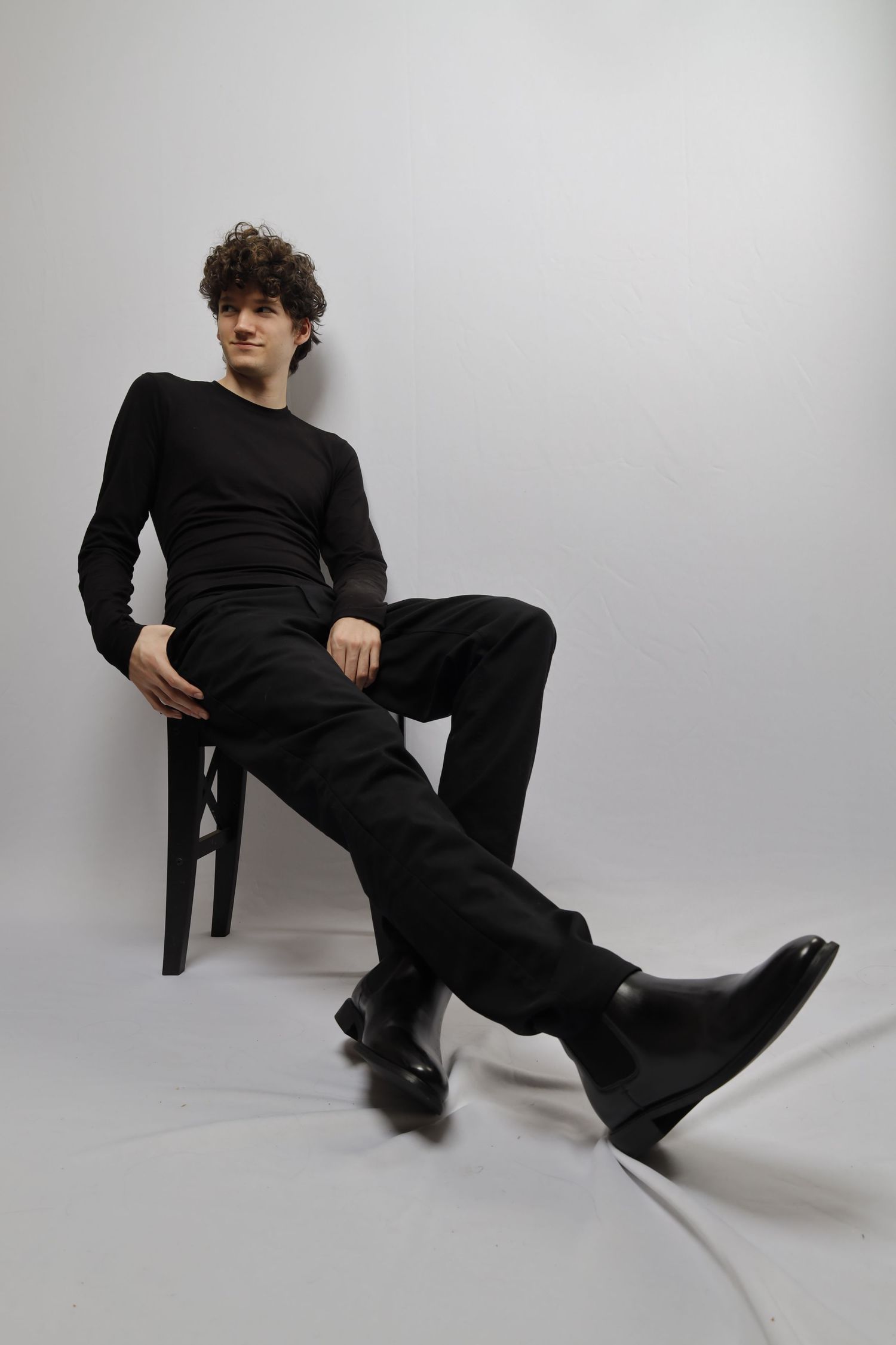 A person in an all-black outfit sits casually on a chair against a light gray background in a minimalist studio setting.