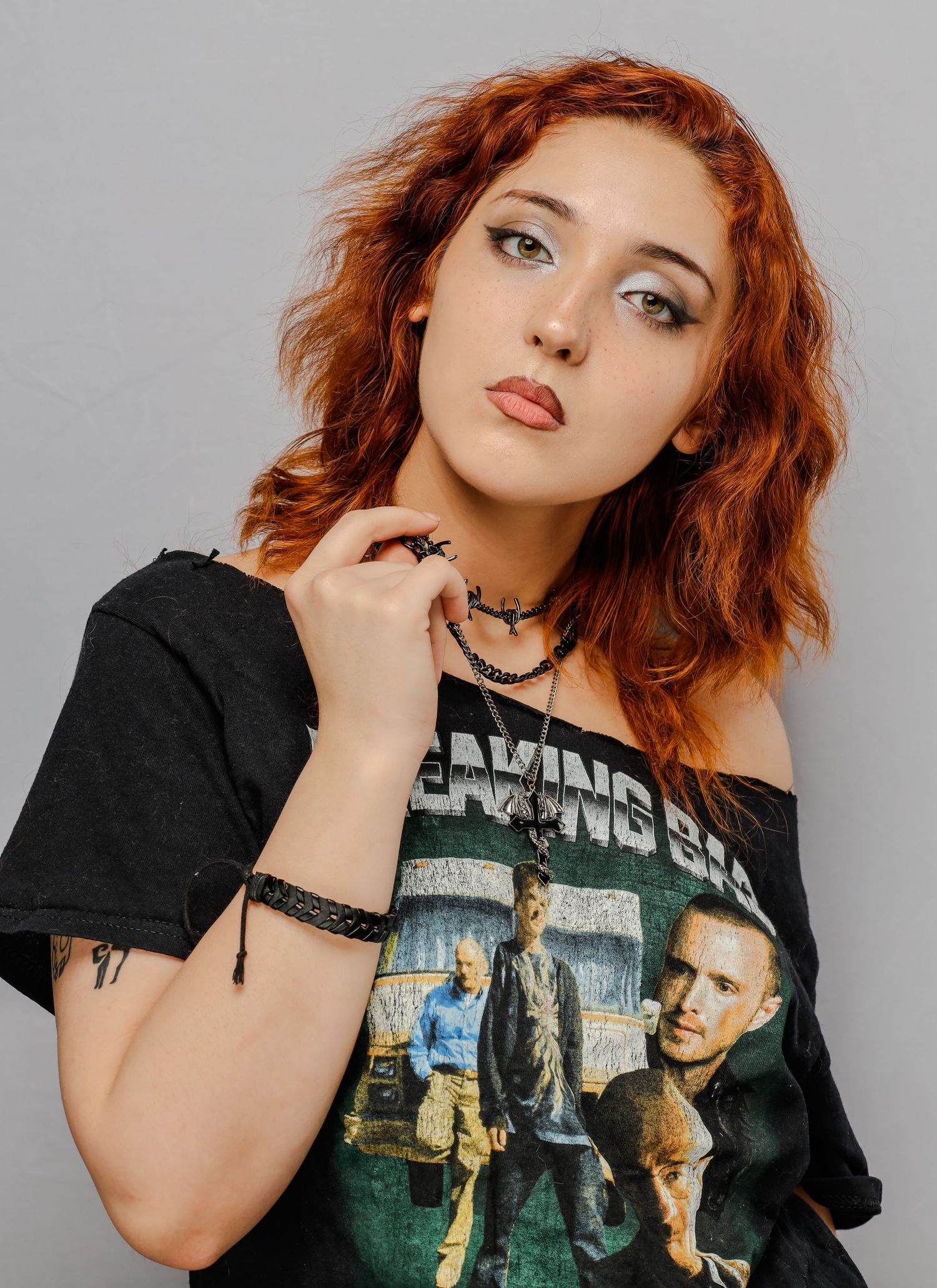 Person with red wavy hair wearing a black band t-shirt and choker necklace poses against grey background.